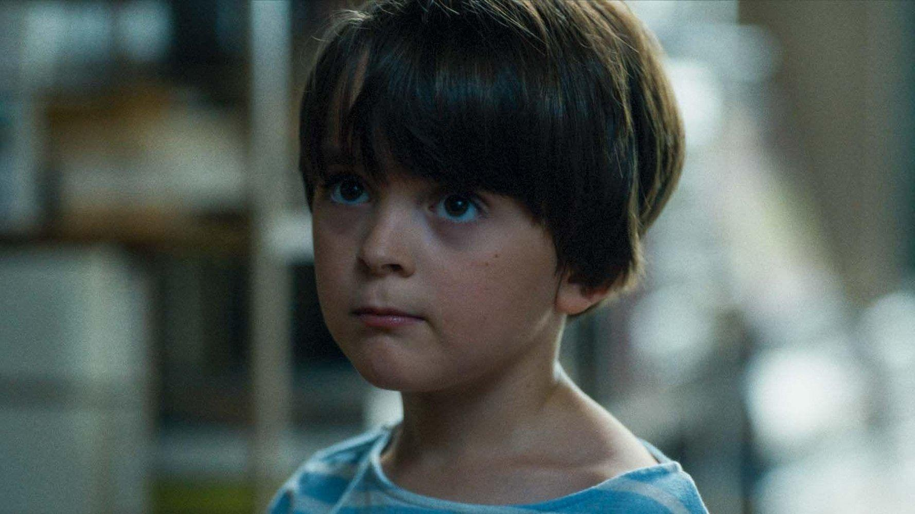 Ein Junge mit dunklem Haar und Augen blickt nach oben. Er trägt ein blau-weiß gestreiftes Hemd. Der Hintergrund ist unscharf.