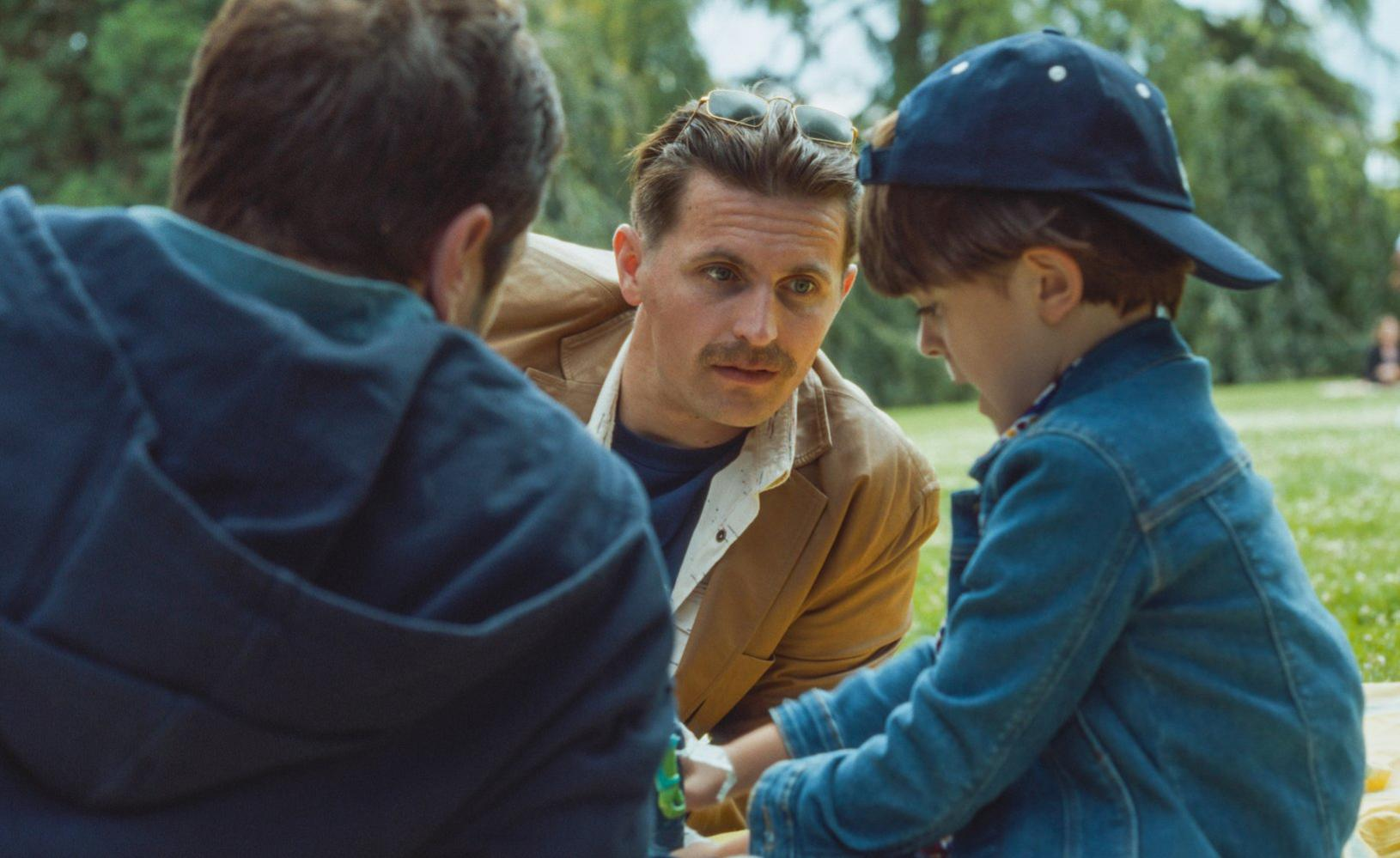 Drei Personen auf einer Picknickdecke. Ein Mann in einer braunen Jacke blickt ein Kind mit blauer Mütze an. Ein weiterer Mann im Vordergrund.