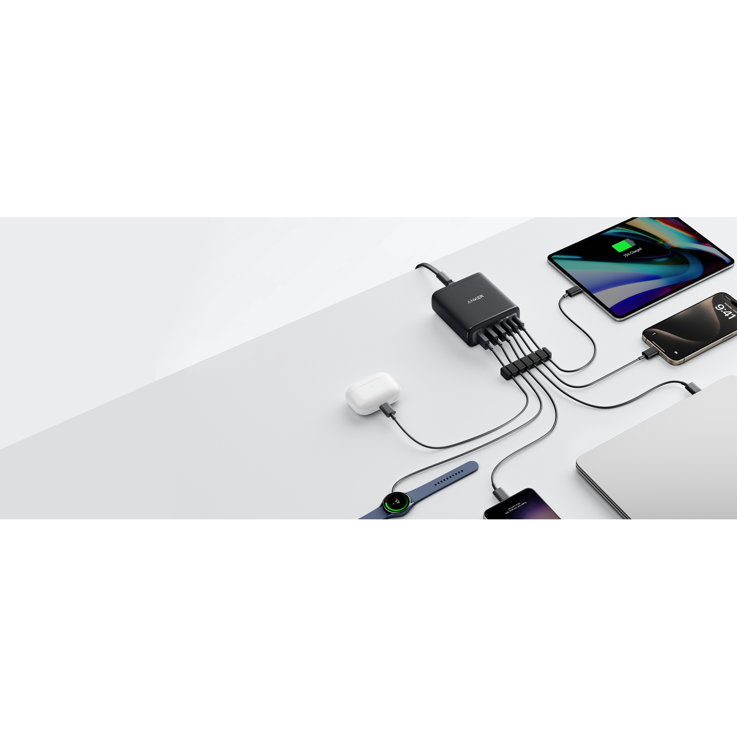 Une station de charge noire Anker est connectée à plusieurs appareils, dont une tablette, un téléphone, une montre, un ordinateur portable et des écouteurs. Tous chargent.