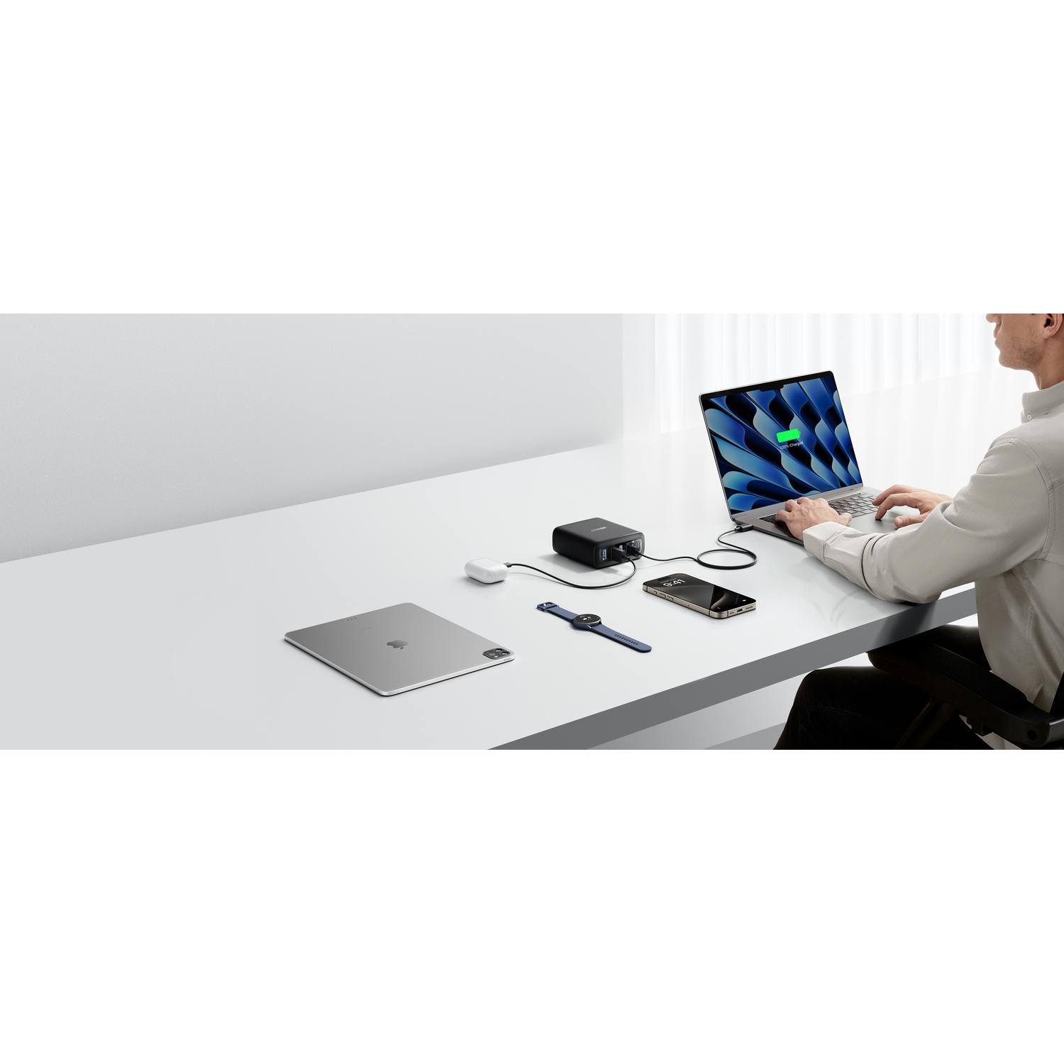 Une personne utilise un ordinateur portable, avec un téléphone, une tablette, une montre connectée et une station de recharge sur un bureau blanc. L'écran montre la charge.