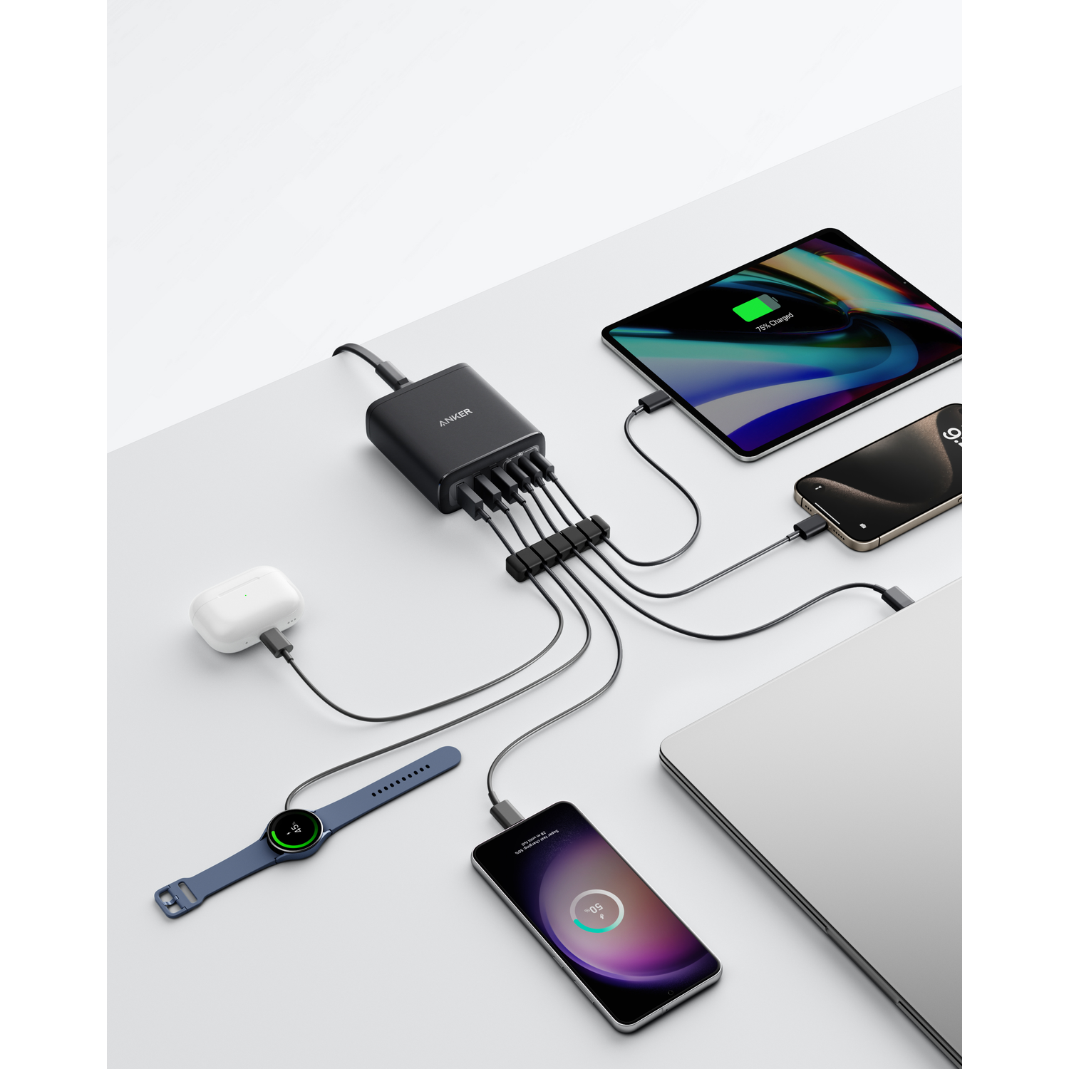 Une station de charge noire Anker charge plusieurs appareils : iPad, téléphone, ordinateur portable, montre, écouteurs et un autre téléphone. Tous les appareils chargent.