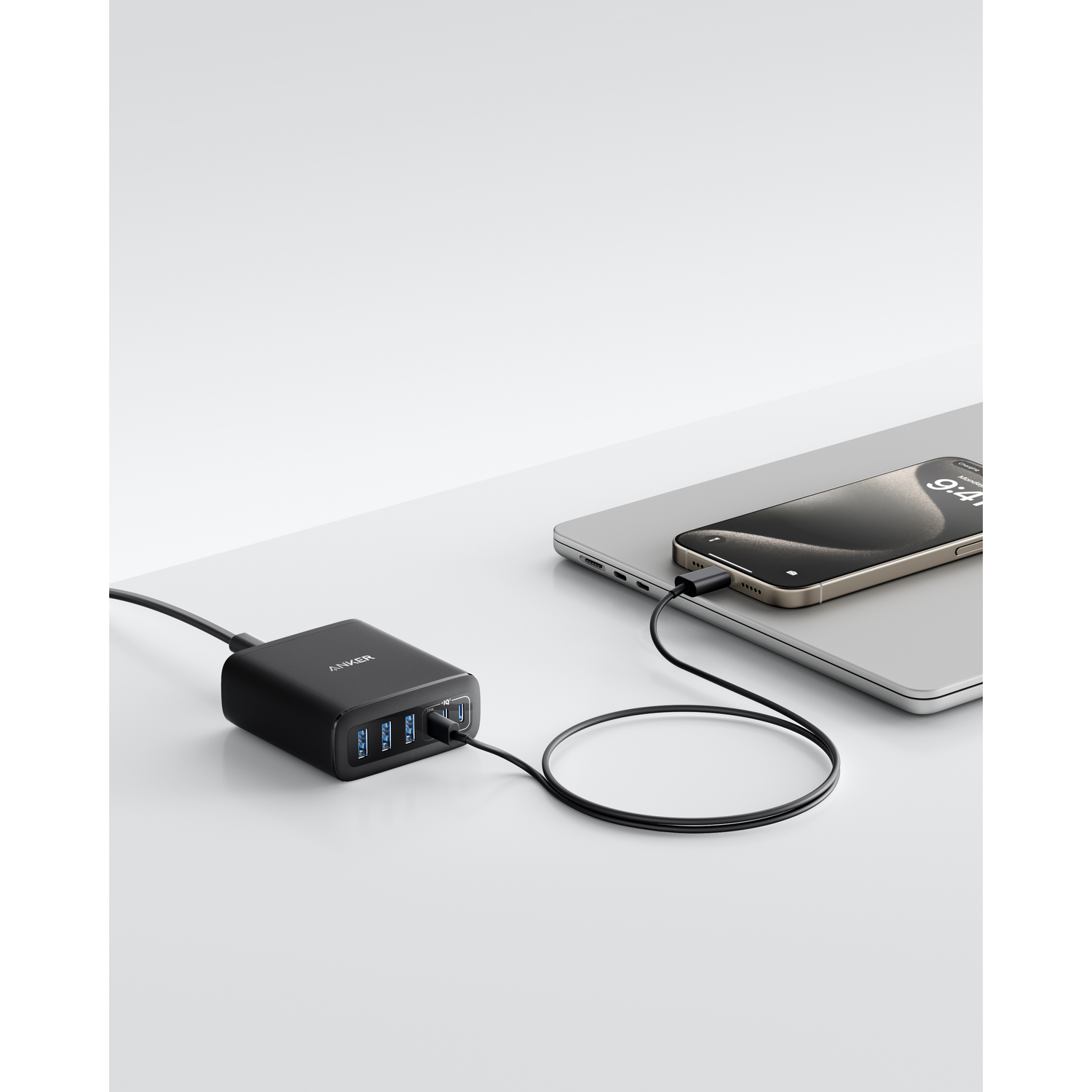 Chargeur Anker noir avec téléphone et ordinateur portable connectés. Le téléphone affiche 9h41. Surface blanche, configuration minimaliste.