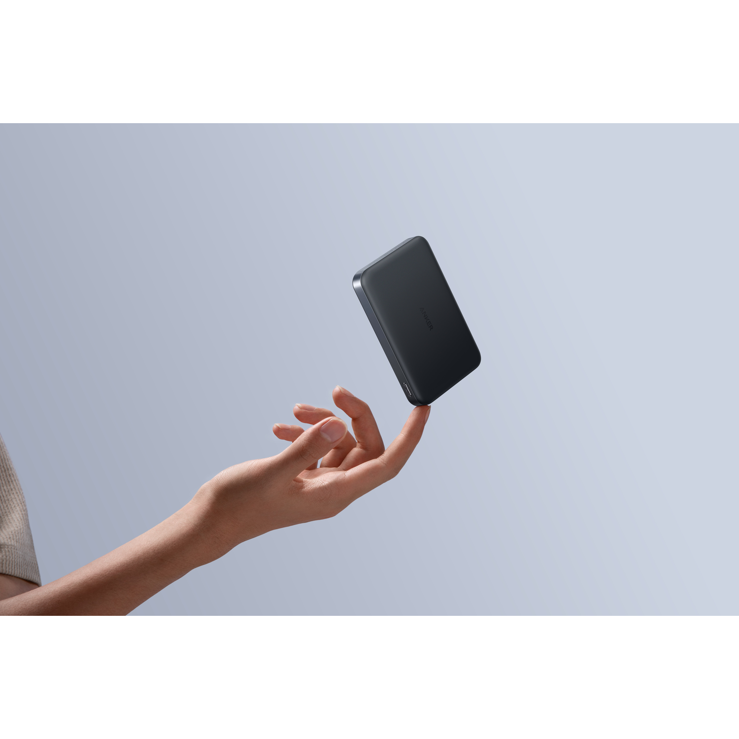 A hand balances a black Anker power bank on a finger. The background is a light blue gradient. The hand is fair-skinned.