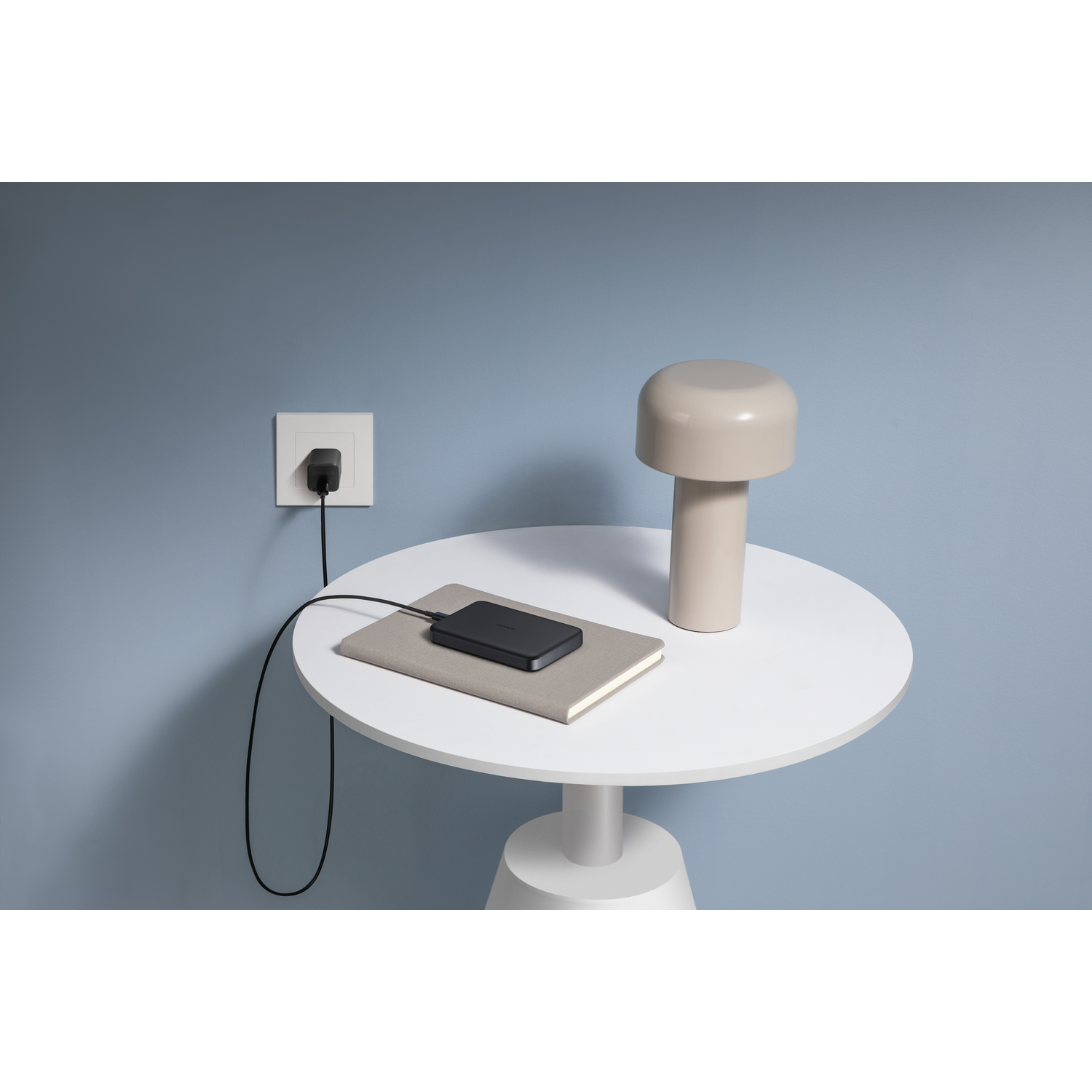 A black charger is plugged into a wall outlet. A notebook, lamp, and charger are on a white table against a blue wall.