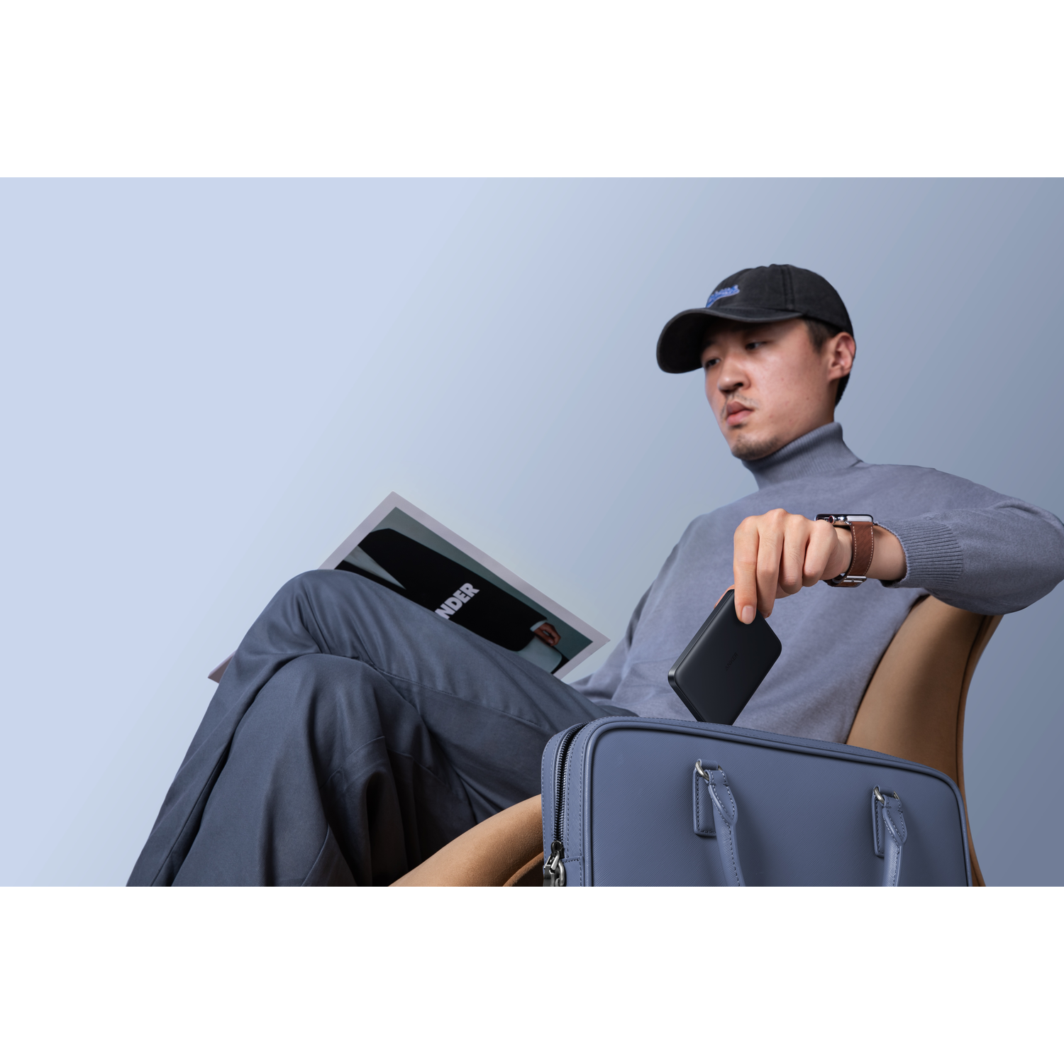 Man in grey turtleneck and cap holding a black device, seated with a magazine and a blue bag.