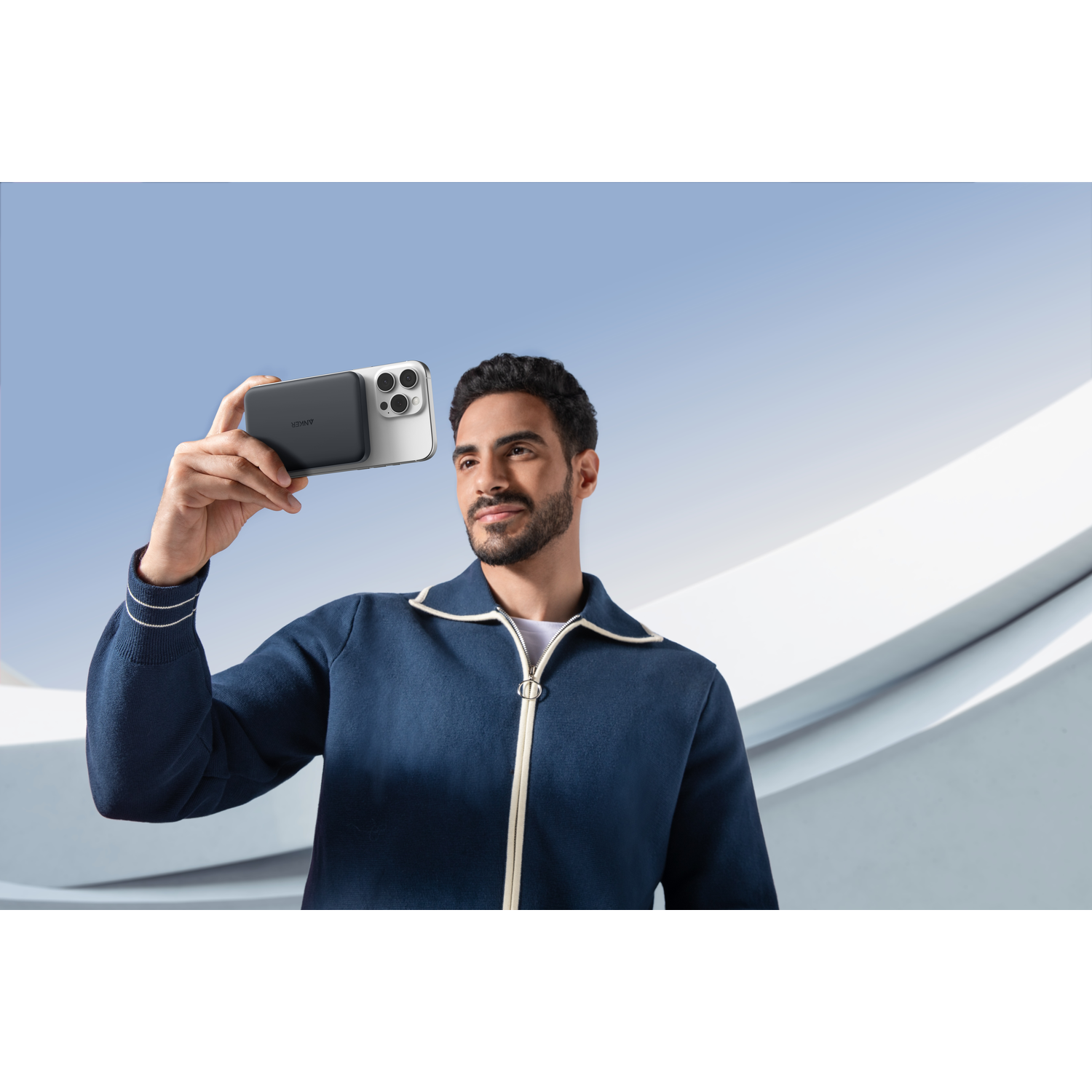 A man holds a phone with a charger. He is wearing a blue jacket. The background is a light blue sky and white structure.