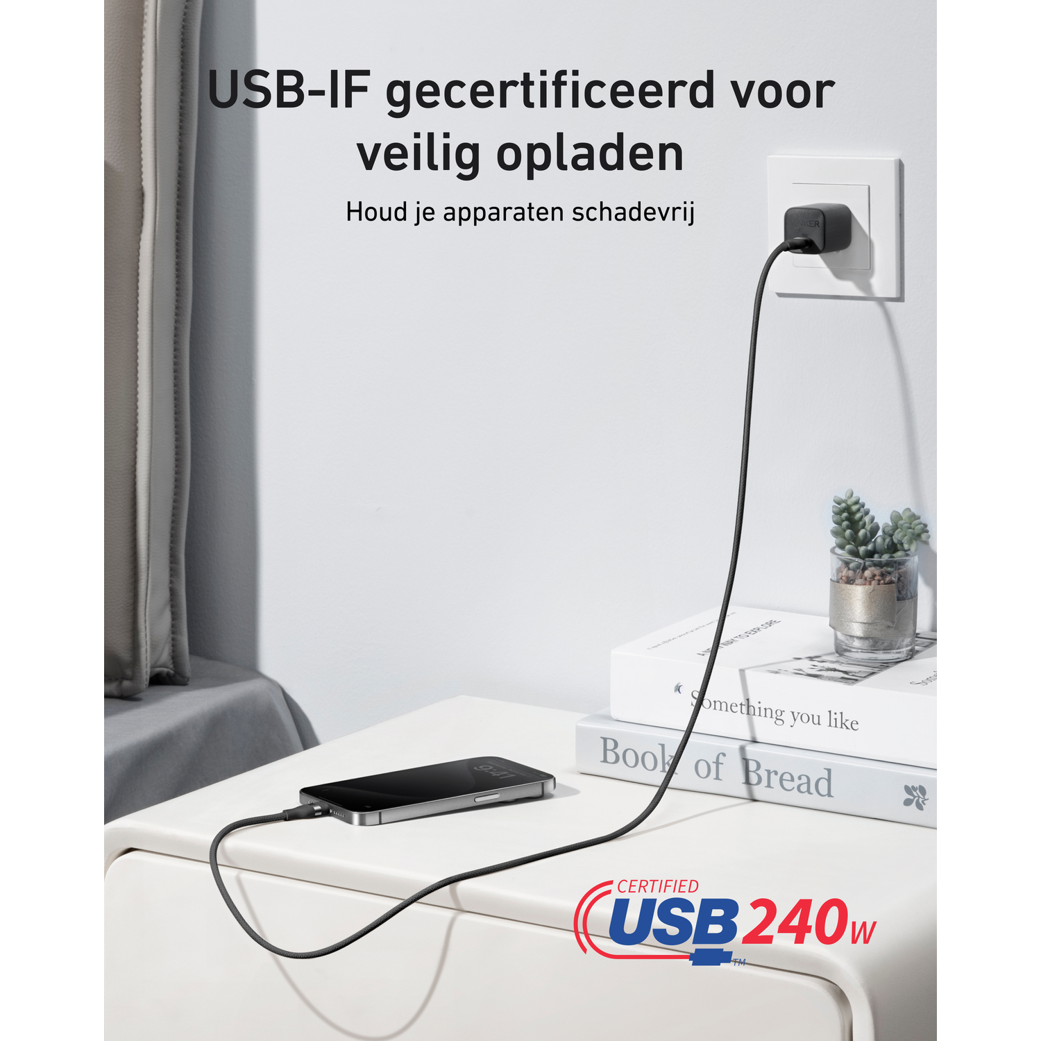 Un téléphone se charge sur une table blanche. Un câble USB se connecte à une prise murale. Livres et plante en arrière-plan.