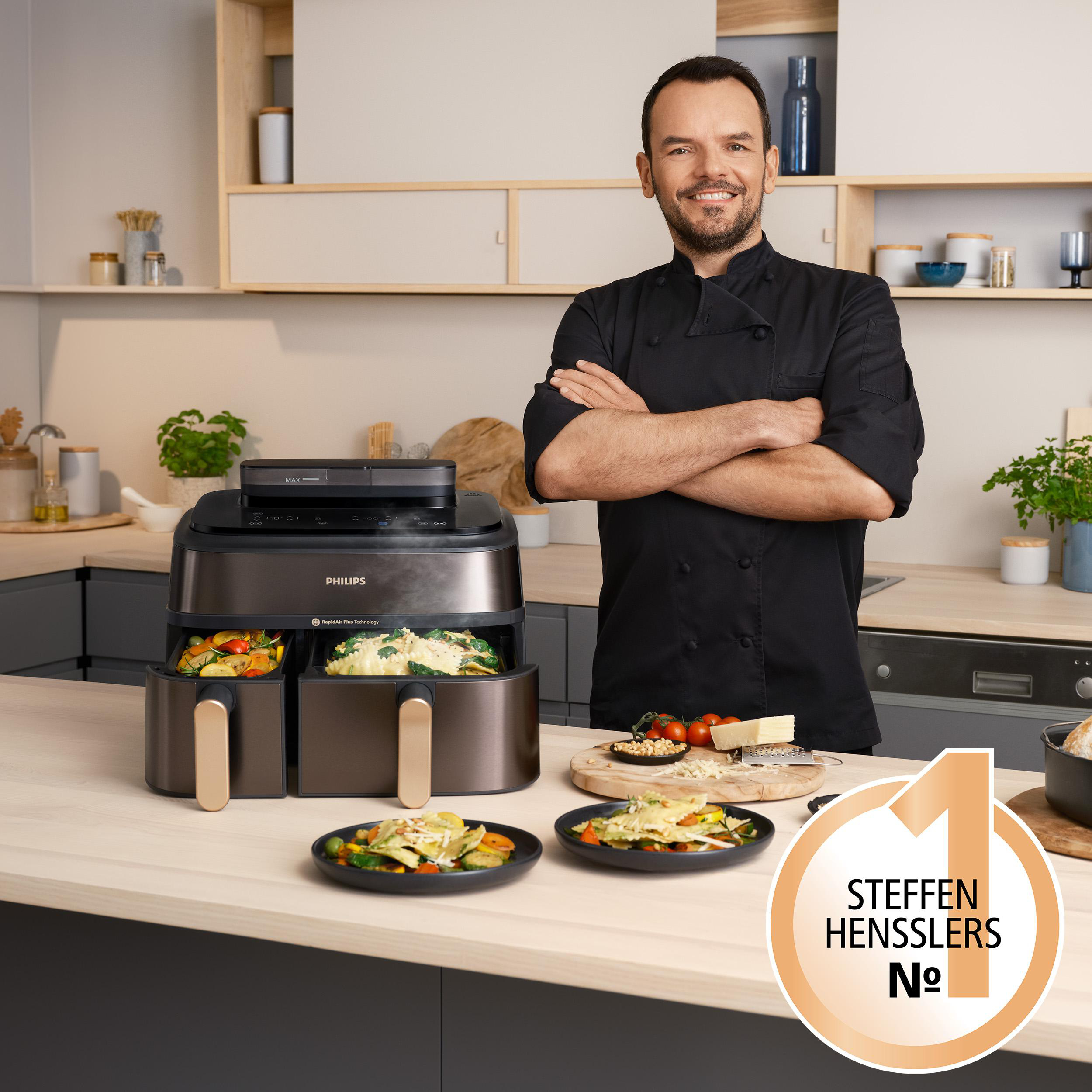 Koch mit verschränkten Armen in der Nähe einer Heißluftfritteuse mit Essen. Teller mit Essen stehen auf dem Tisch. Ein Logo befindet sich in der rechten Ecke.