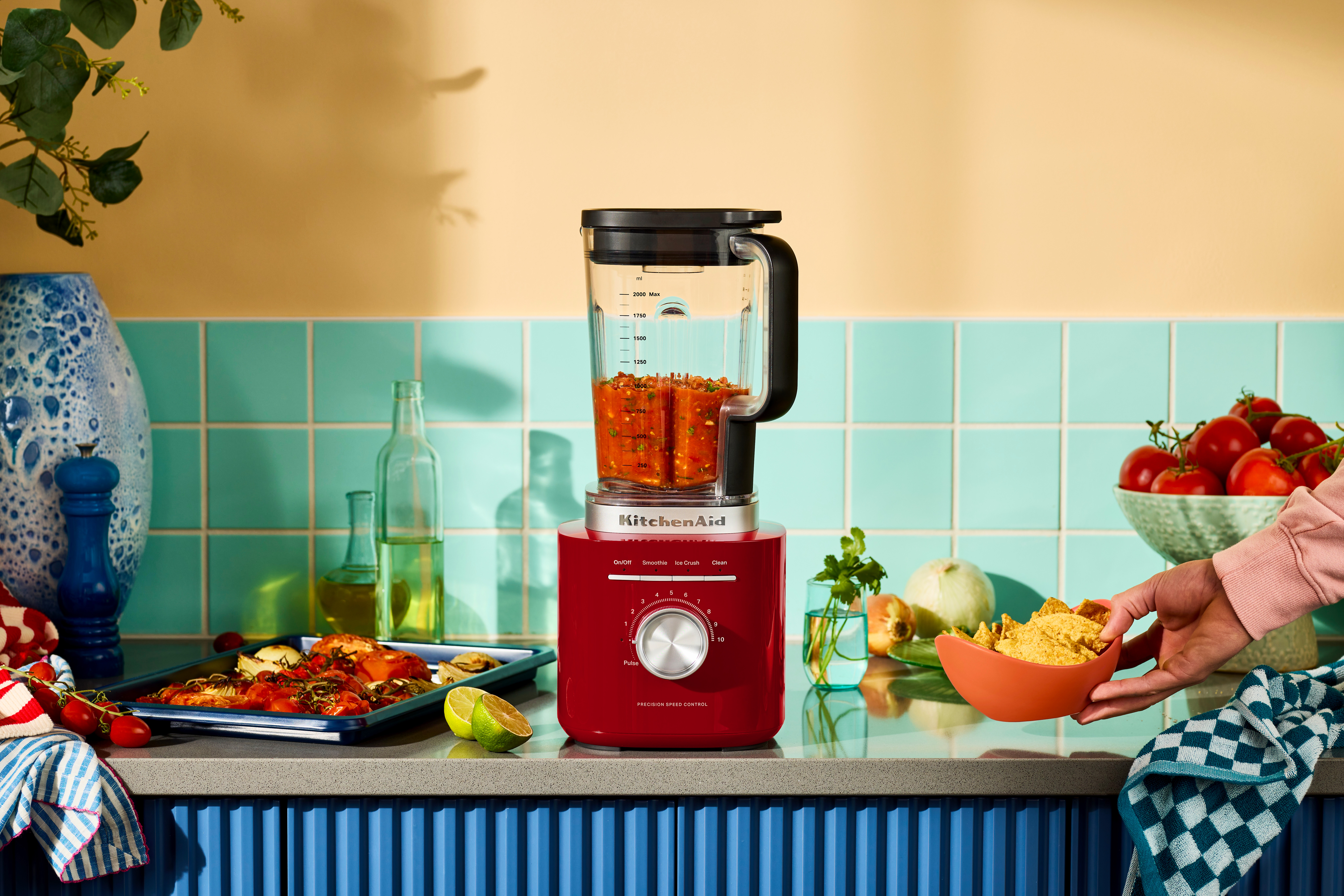 Ein roter Mixer mit Salsa steht auf einer Theke. Hand greift nach Chips. Tomaten, Limette und ein Tablett mit Essen sind sichtbar.