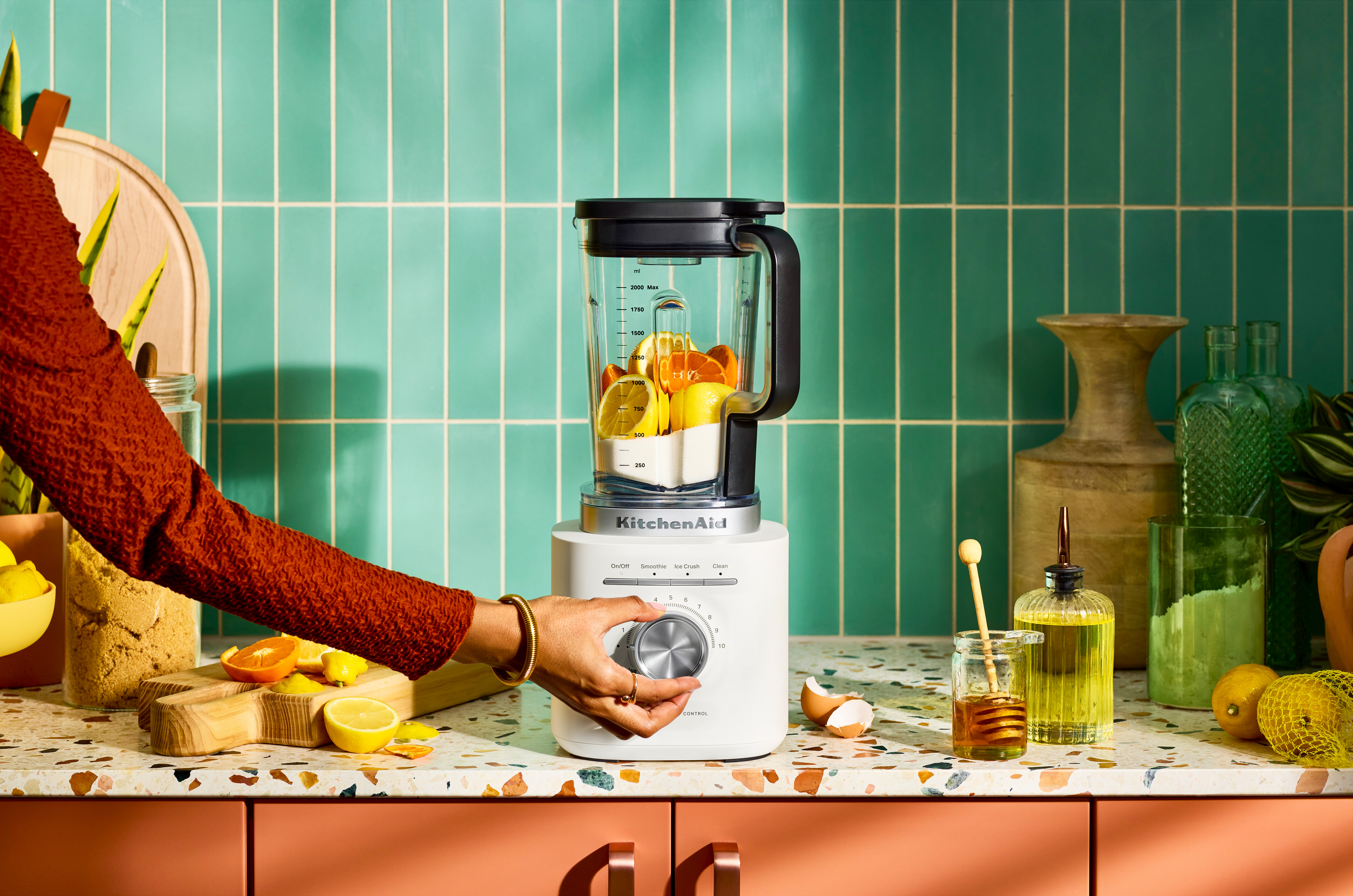 Eine Hand dreht einen Drehknopf des Mixers. Der Mixer enthält Früchte. Der Hintergrund ist eine türkisfarbene Kachelwand.