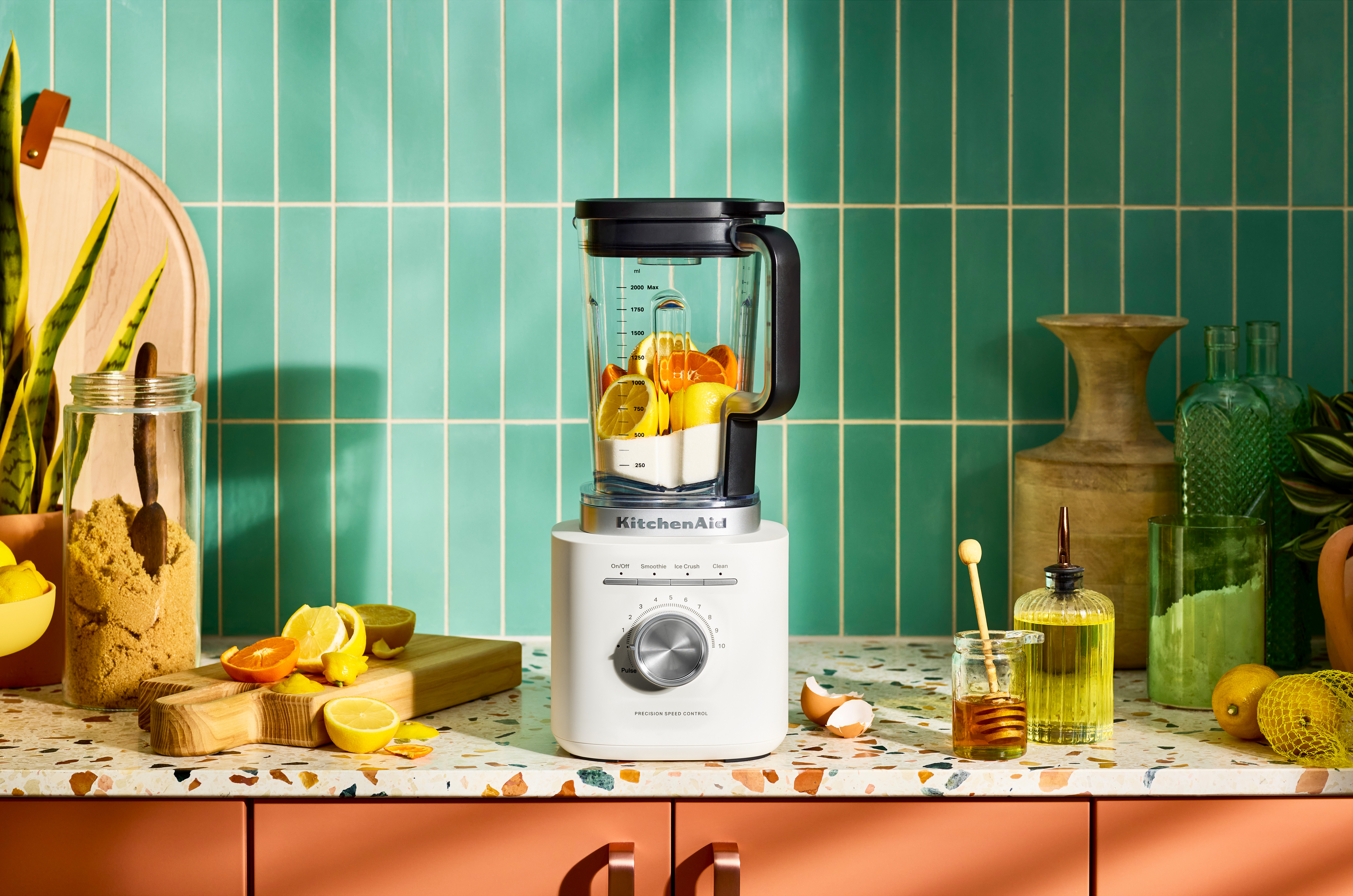 Ein weißer KitchenAid-Mixer mit Zitrusfrüchten im Inneren. Zitronen und Orangen auf einem Schneidebrett. Honig und Öl.
