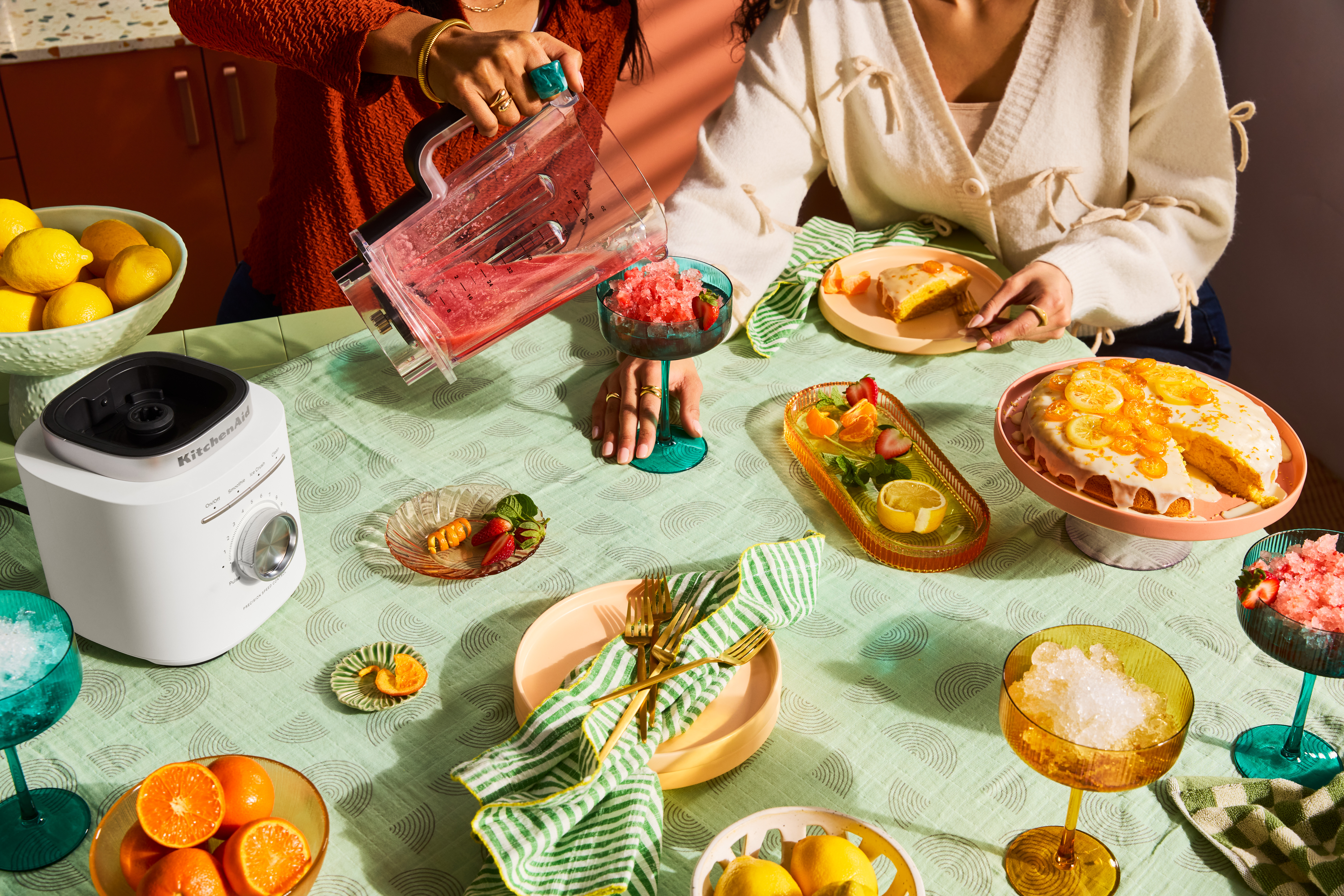 Zwei Personen bereiten eine Mahlzeit zu. Ein Mixer gießt rosa Flüssigkeit in ein Glas. Ein Kuchen und Obst stehen auf dem Tisch.