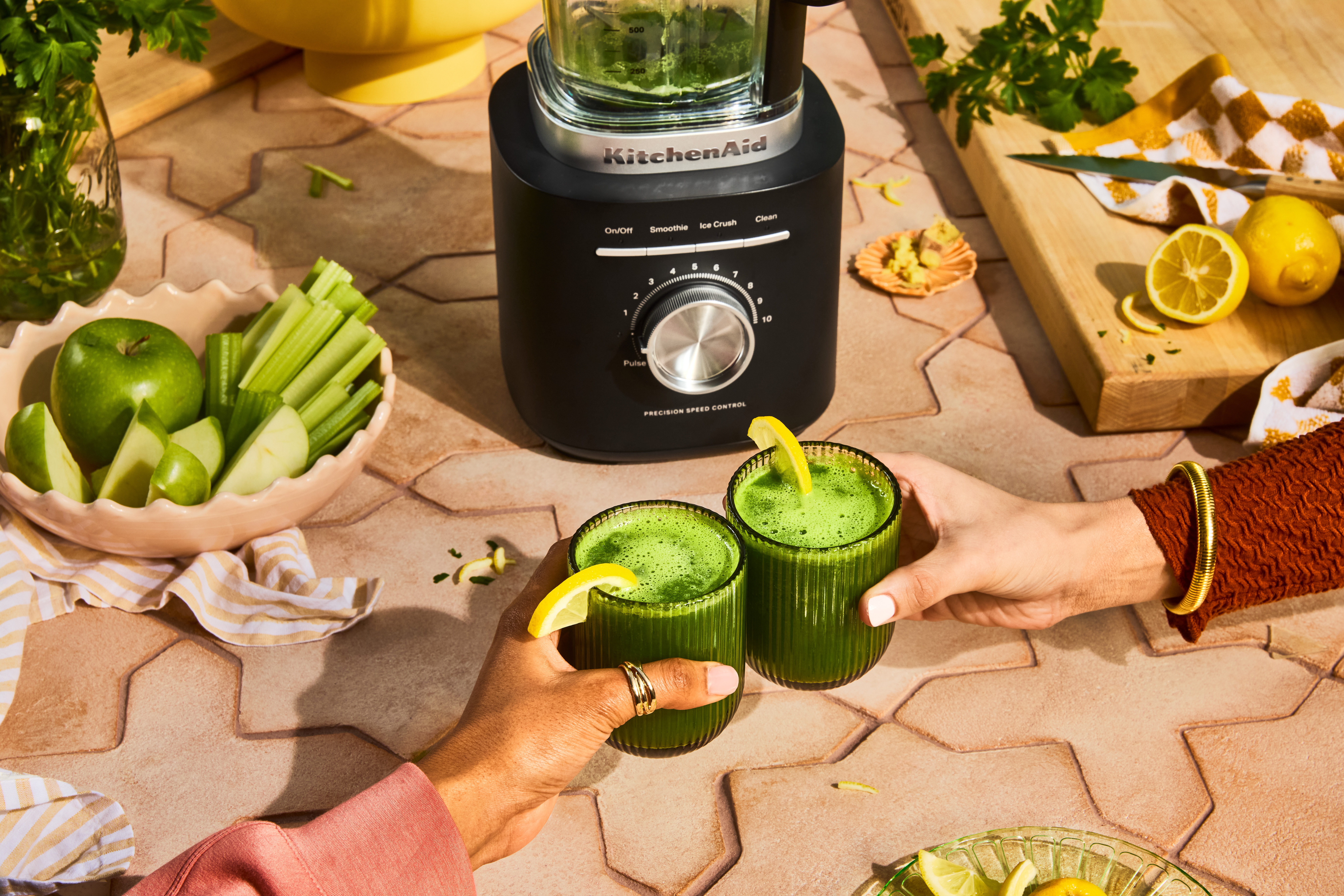 Grüne Smoothie-Gläser werden hochgehalten, Zitronen, Äpfel und ein Mixer stehen auf dem Tisch.