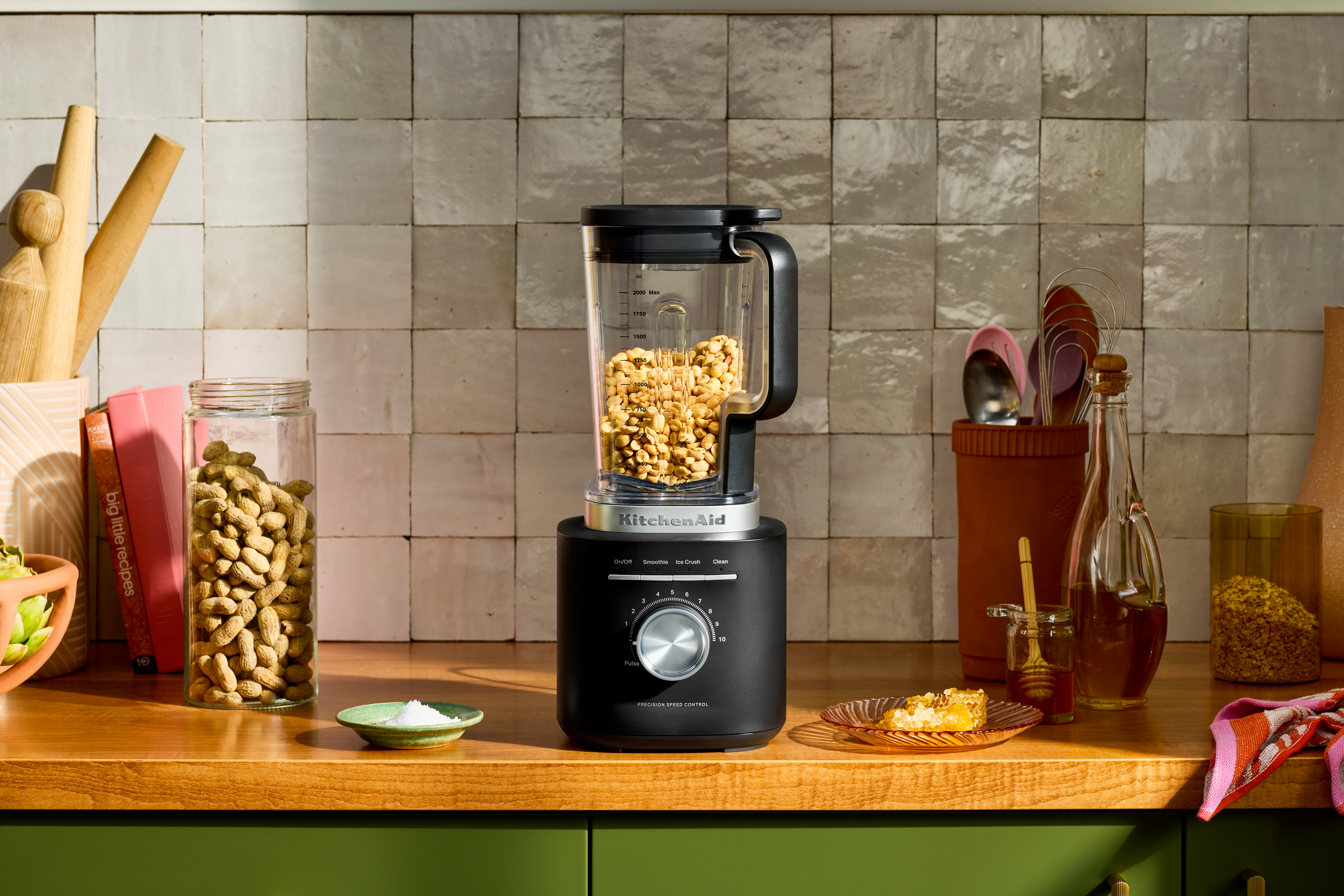 Eine Küchenszene mit einem Mixer, gefüllt mit Erdnüssen, Honig, Salz und anderen Zutaten auf einer Holztheke.
