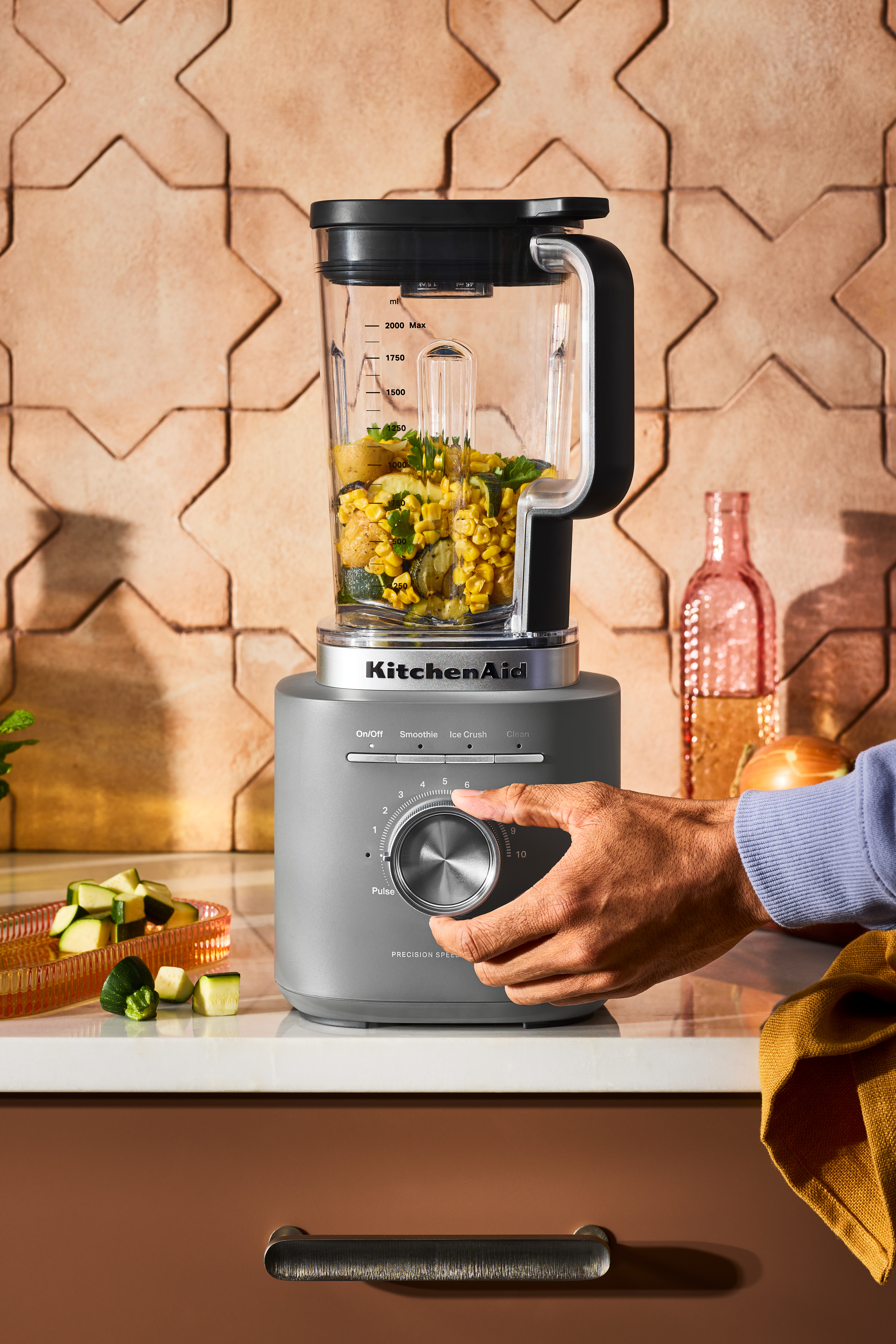 Ein KitchenAid-Mixer mit Gemüse. Eine Hand dreht den Geschwindigkeitsregler. Hintergrund: geflieste Wand und rosa Glasflasche.