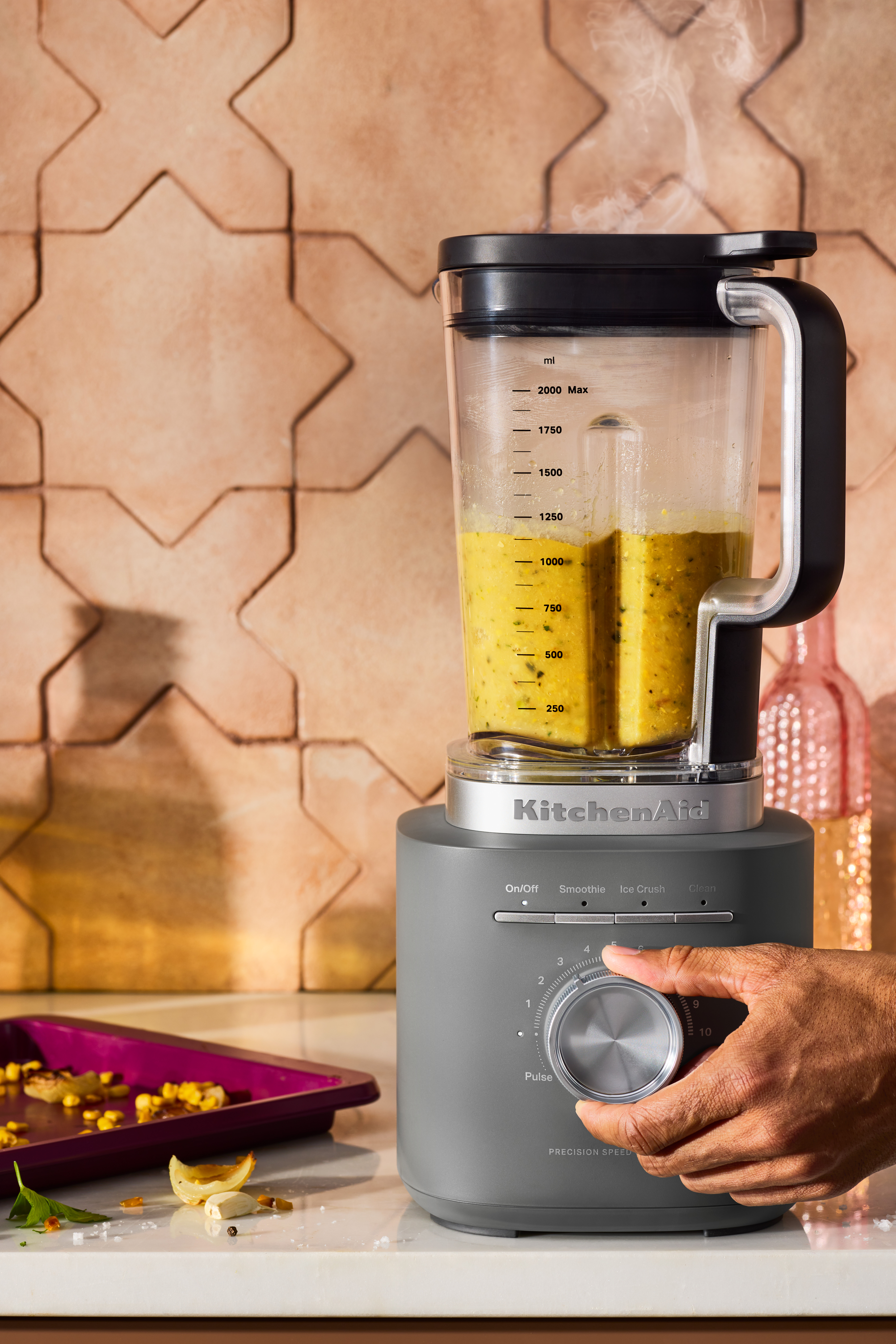 Ein grauer KitchenAid-Mixer mit gelber Flüssigkeit. Eine Hand dreht den Geschwindigkeitsregler. Zutaten liegen auf der Theke.