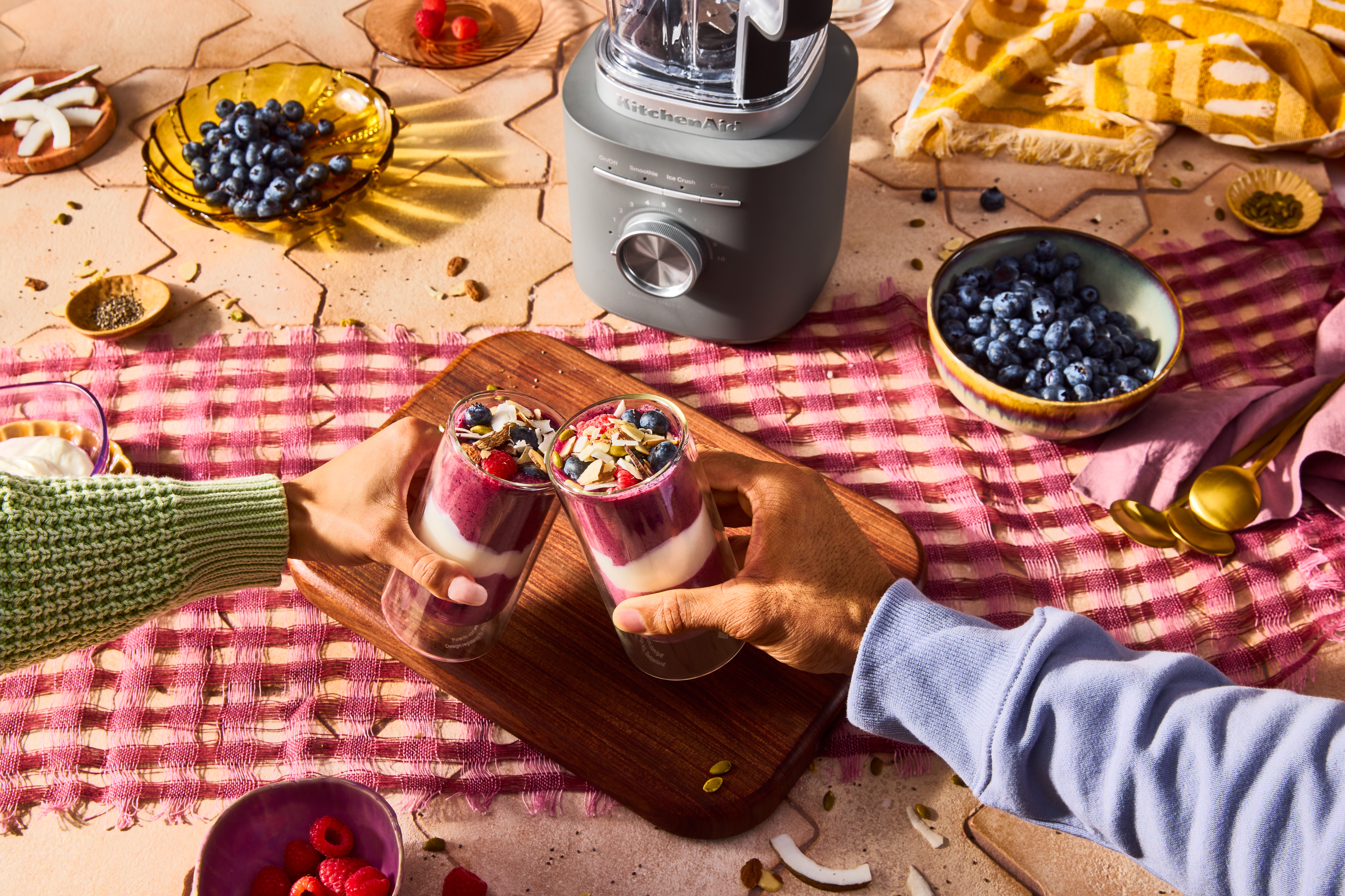Zwei Hände halten Smoothie-Gläser. Zutaten sind Beeren, Kokosnuss und Samen. Im Hintergrund ein KitchenAid-Mixer.