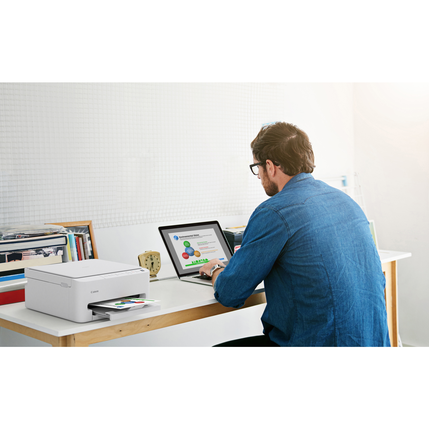 A man in a blue shirt works on a laptop. A white printer sits on the desk, with a printout. A clock and books are on the desk.