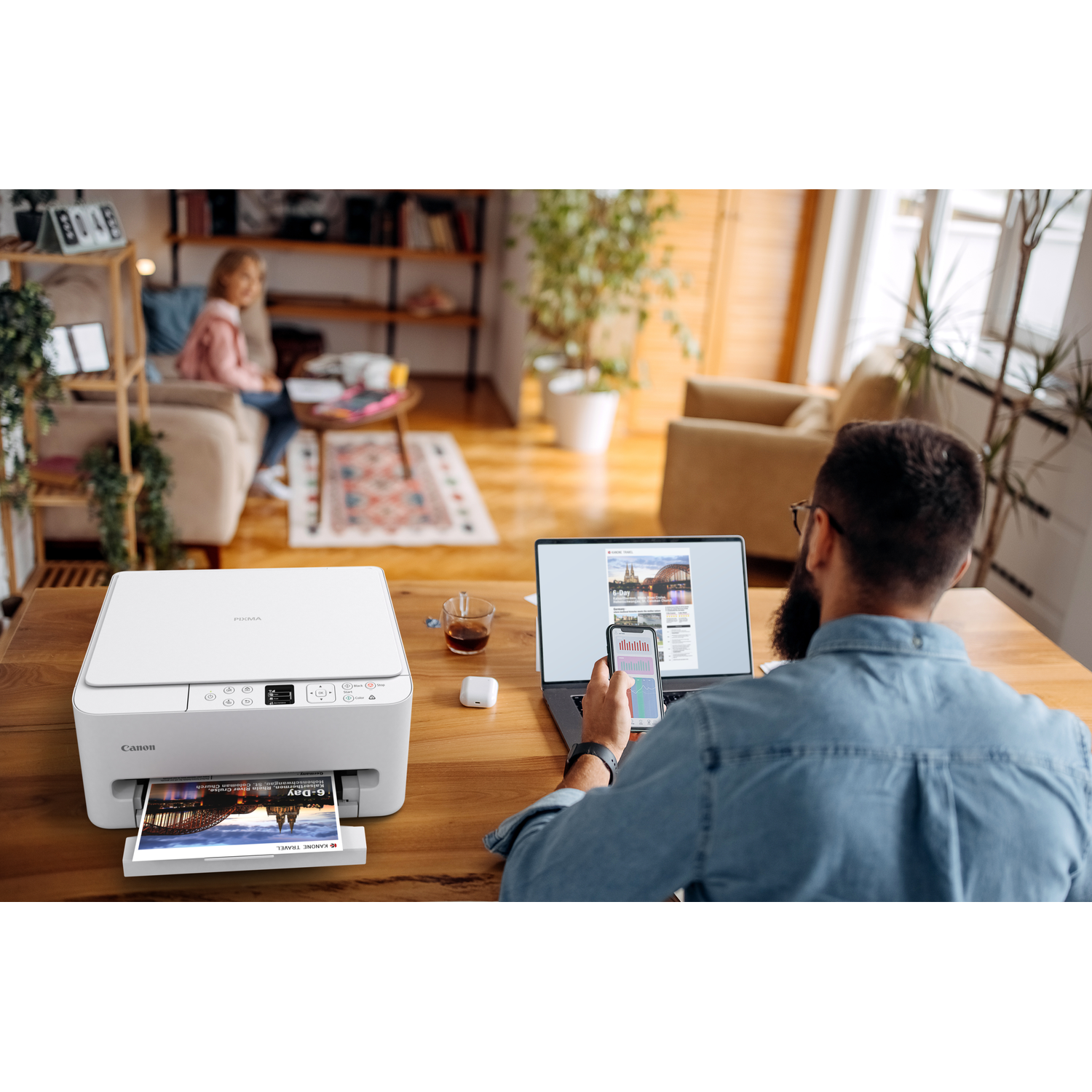 A man prints from a laptop and phone. A white printer sits on a wooden table, with a printed page. A woman sits on a sofa.