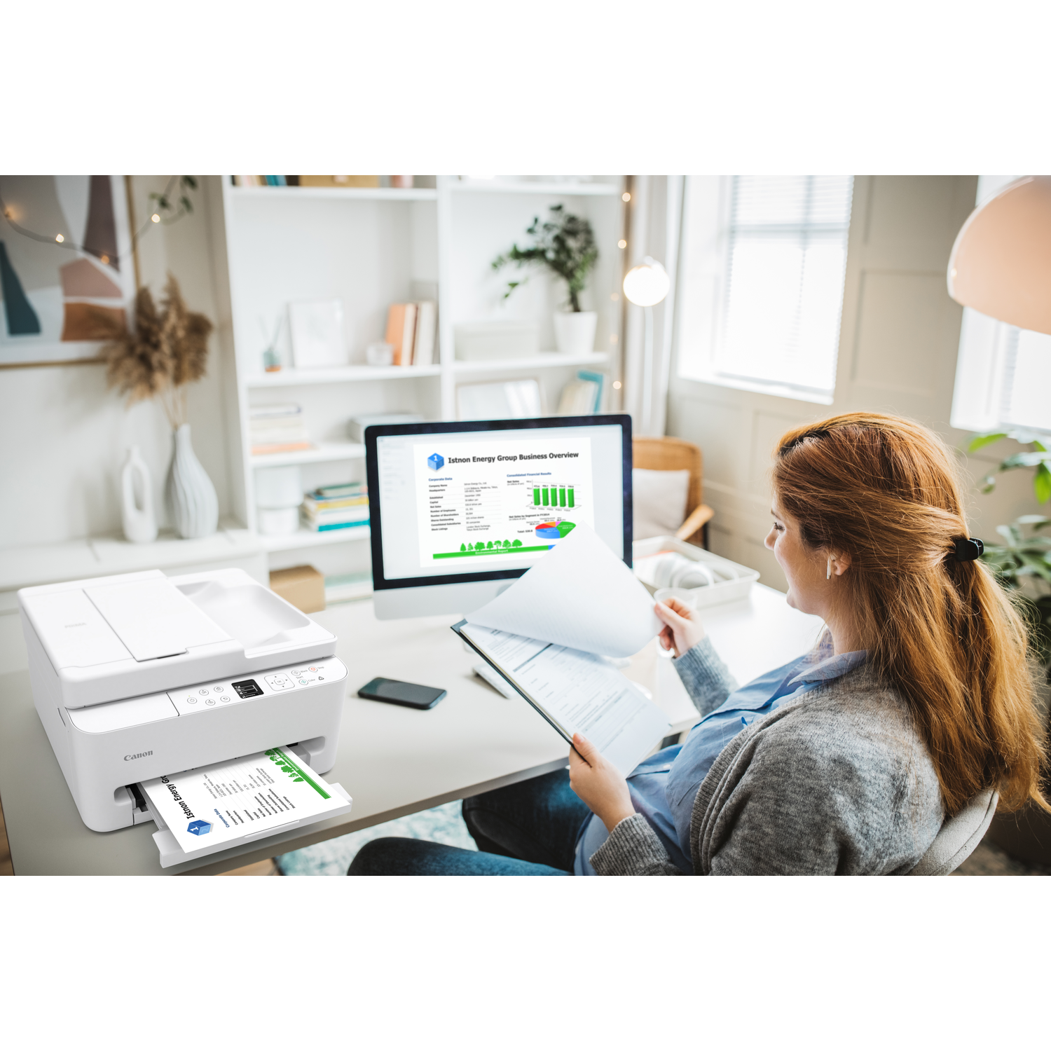 A woman reviews papers at a desk with a printer and computer. The screen shows a business overview.