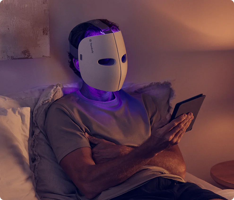 Eine Person mit weißer Gesichtsmaske benutzt ein Tablet im Bett. Die Szene wird von violettem Licht beleuchtet.