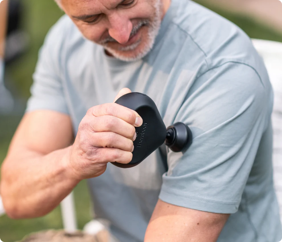 Ein Mann benutzt eine schwarze Massagepistole an seinem Arm. Er trägt ein graues Hemd und lächelt. Der Hintergrund ist grün verschwommen.