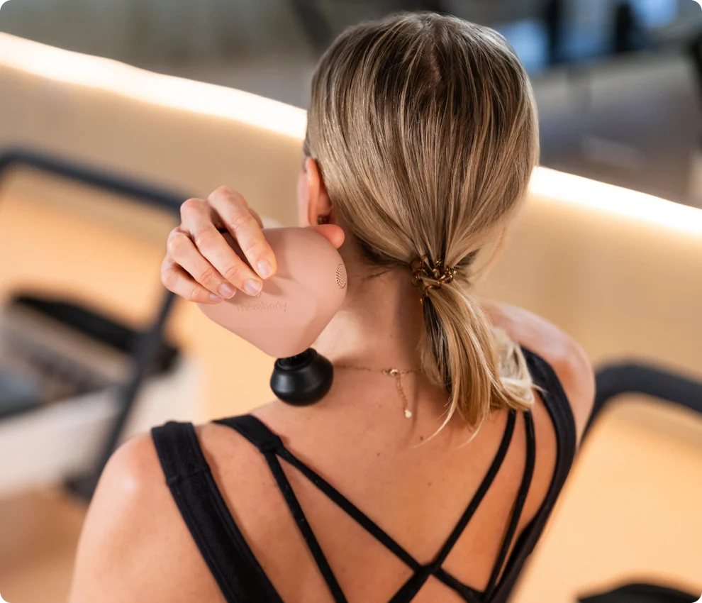 Eine Frau benutzt ein Massagegerät an ihrem Hals. Sie trägt ein schwarzes Oberteil und hat blonde Haare im Pferdeschwanz. Das Gerät ist rosa und schwarz.