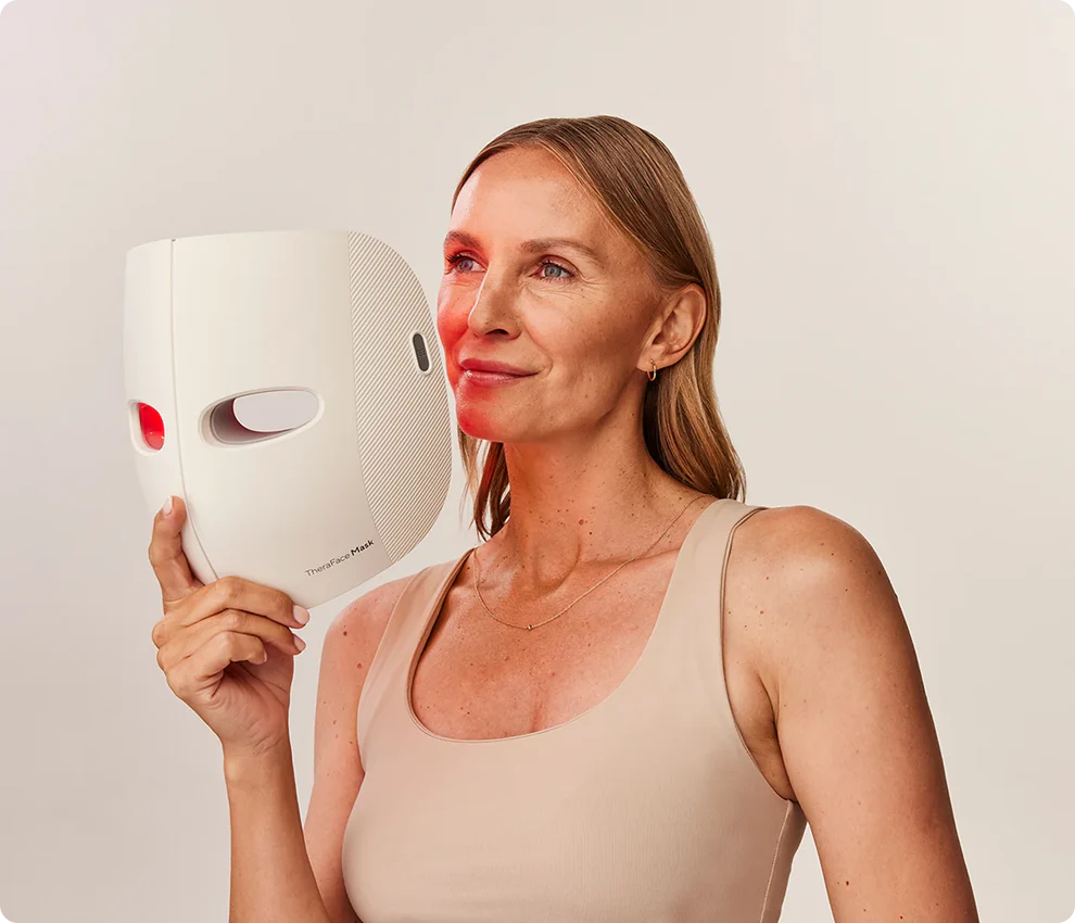 Frau hält eine weiße Gesichtsmaske. Ihr Gesicht wird von rotem Licht beleuchtet. Sie trägt ein beiges Tanktop und eine goldene Halskette.