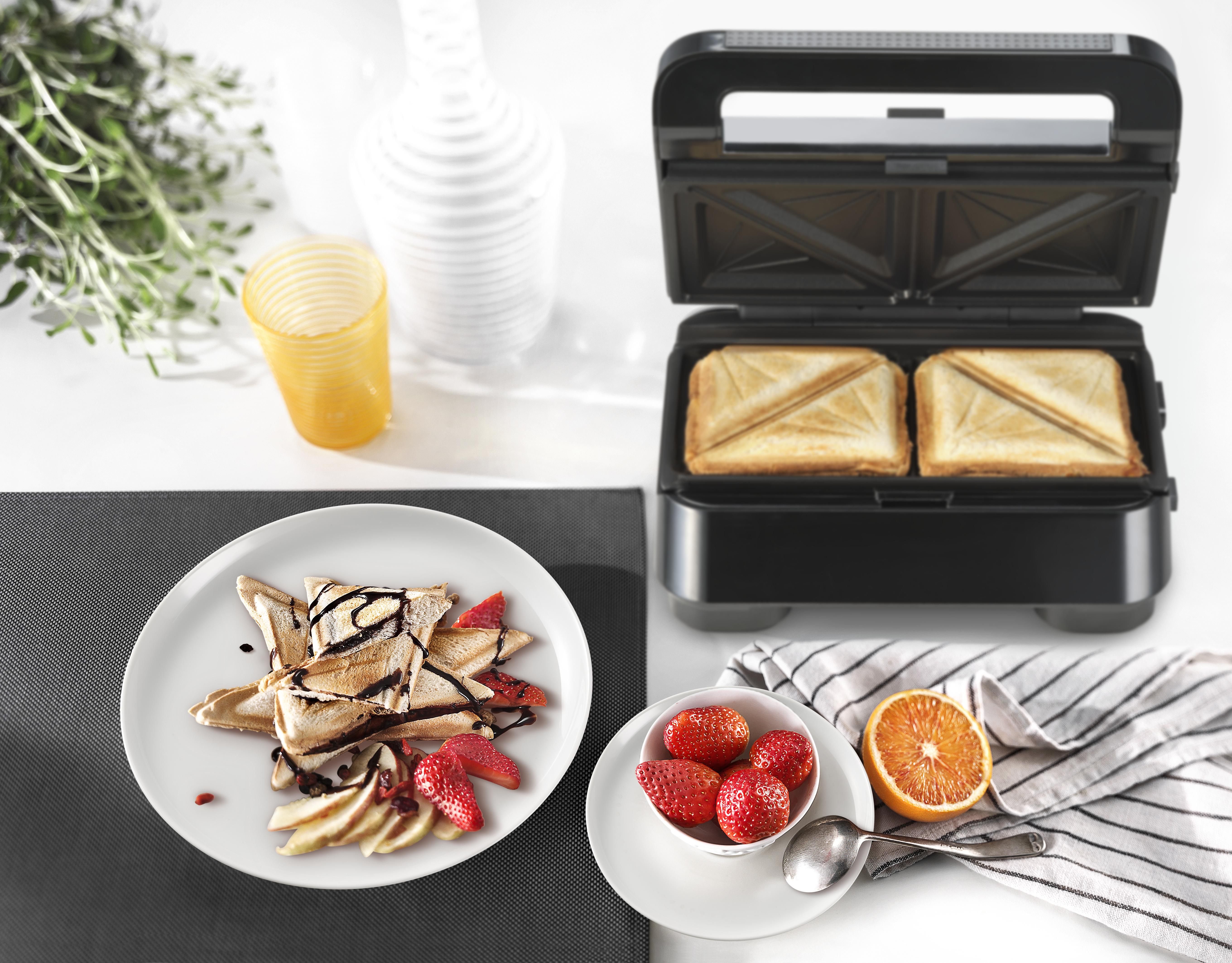 Frühstücksszene mit Sandwichmaker, Sandwiches, Obst und Getränk. Das Essen ist auf einer weißen Oberfläche angerichtet.