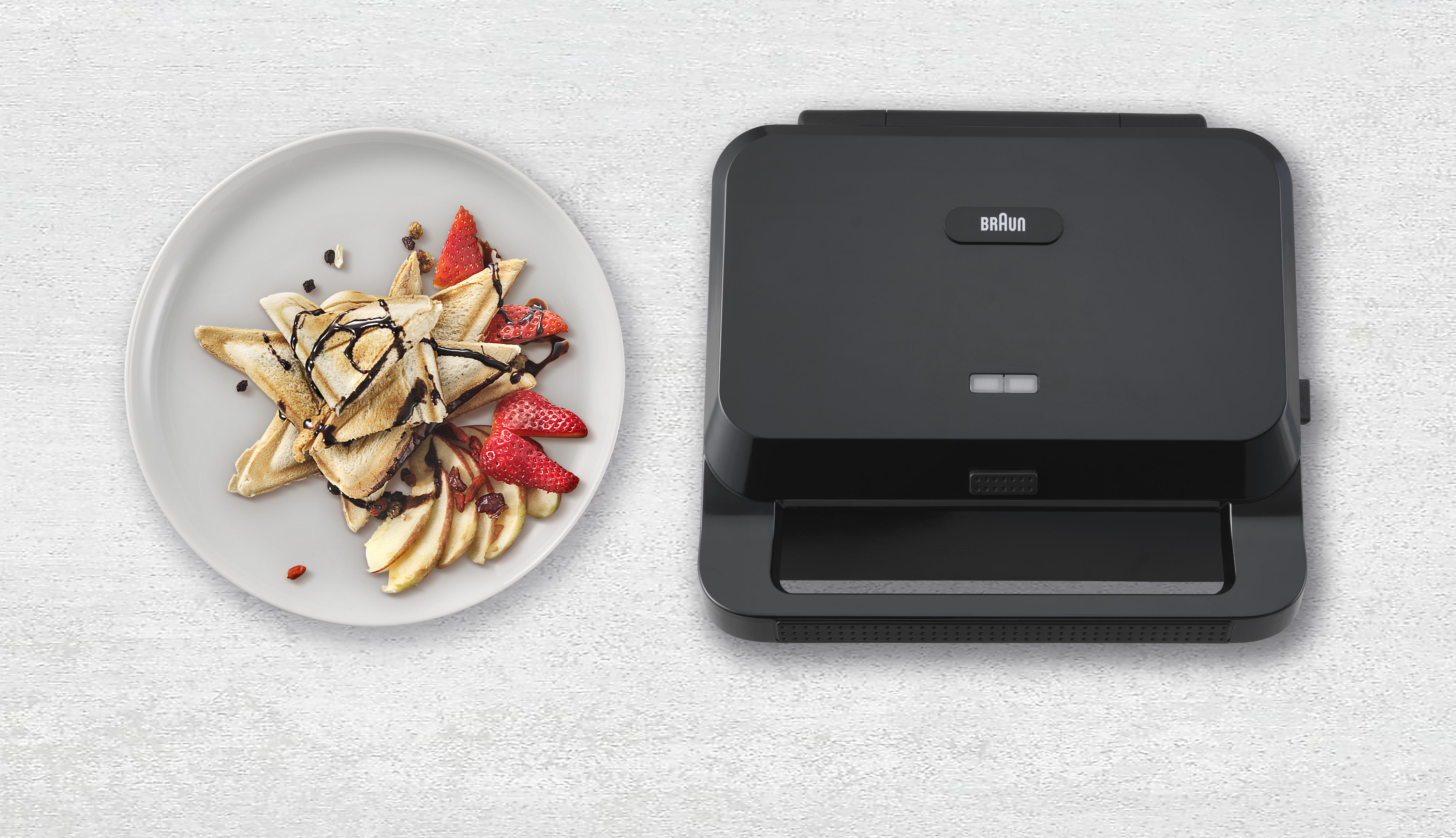 Ein Teller mit Waffeln, Erdbeeren und Äpfeln neben einem schwarzen Braun-Waffeleisen auf weißem Untergrund.