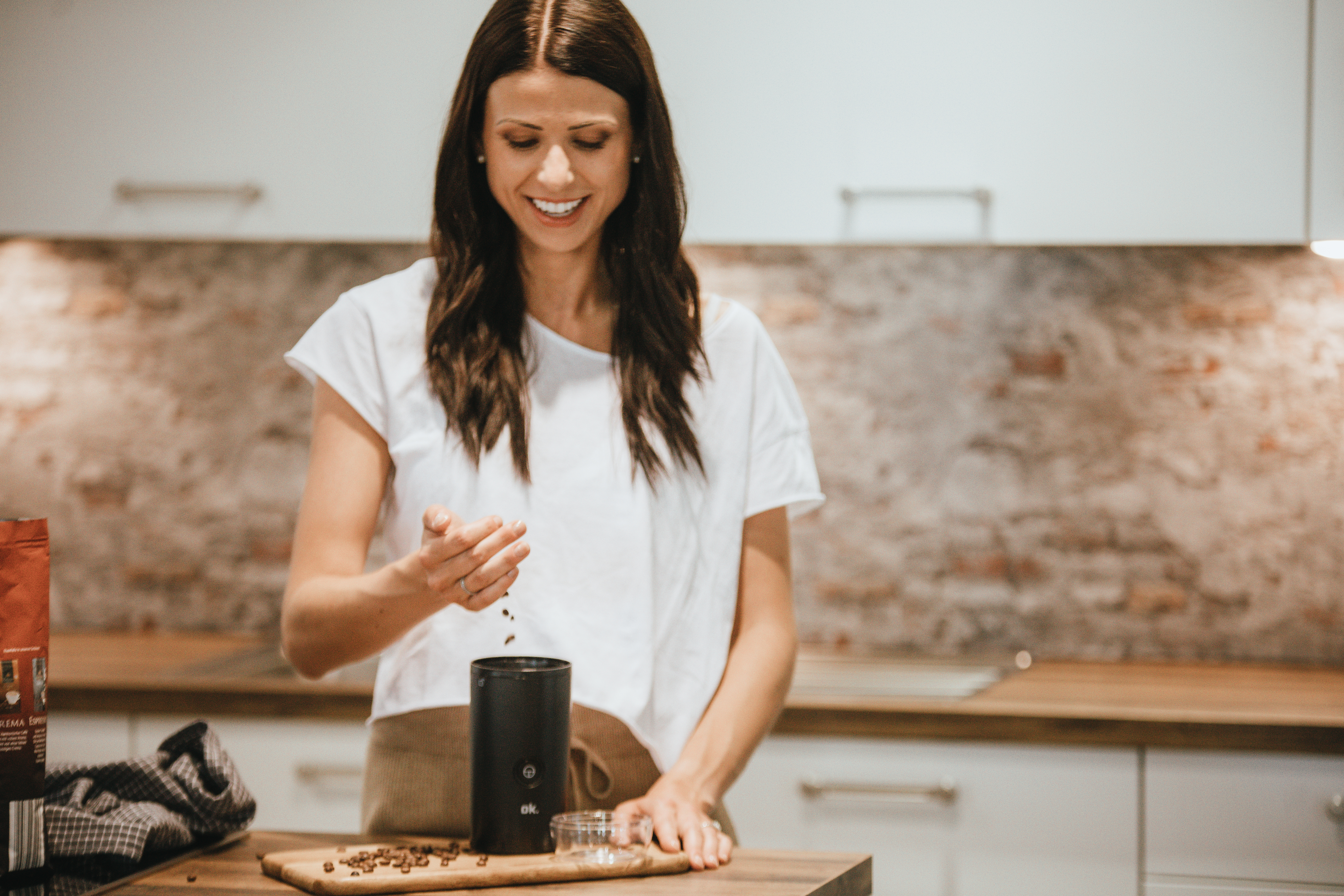 Kobieta uśmiecha się, wsypując ziarna kawy do młynka. Otoczenie kuchni z drewnianym blatem i ceglaną ścianą. Widoczna paczka kawy.