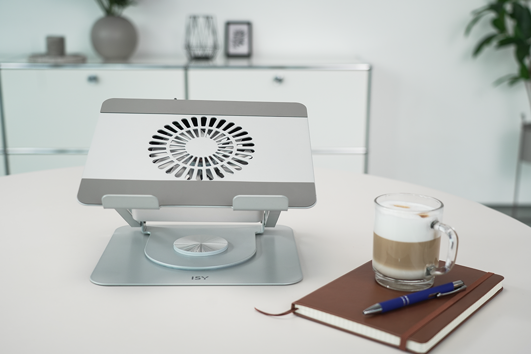 Egy laptop állvány ventilátorral, kávé tollal és jegyzetfüzettel egy fehér asztalon. Háttér: szekrény és növények.