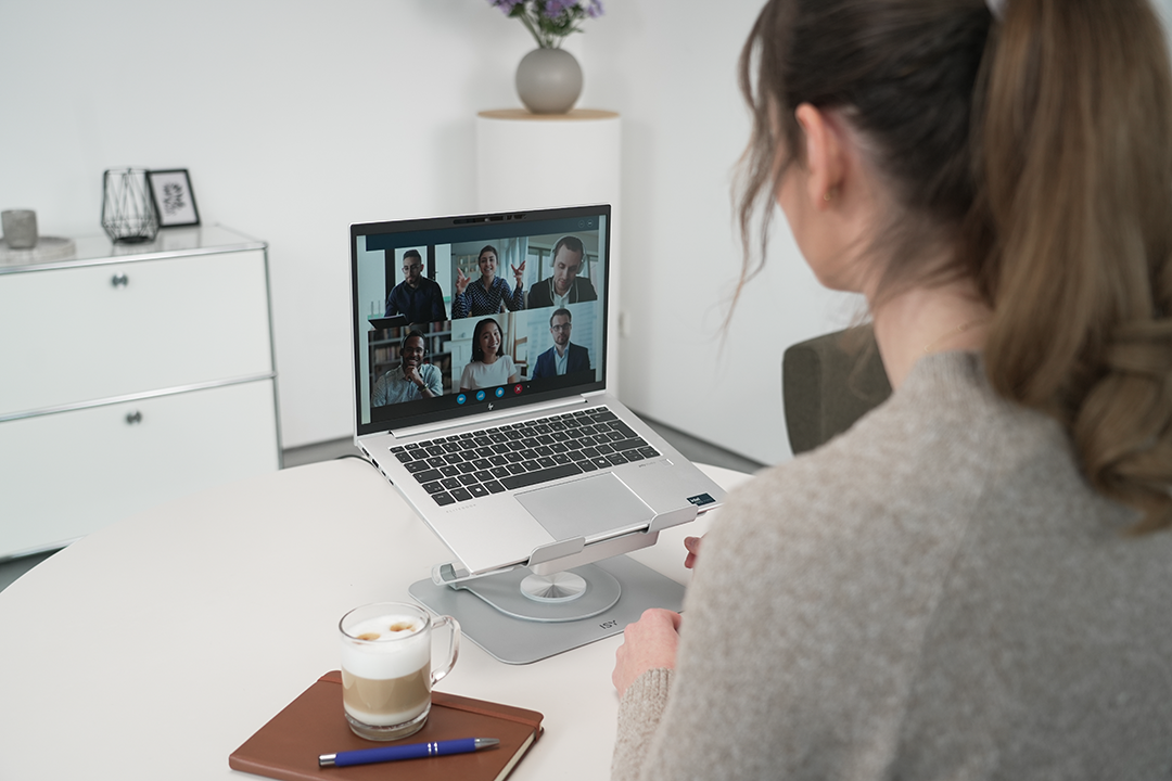 Egy nő videohívást folytat laptopon. Kávé és jegyzetfüzet az asztalon. Fehér szekrények és egy váza a háttérben.