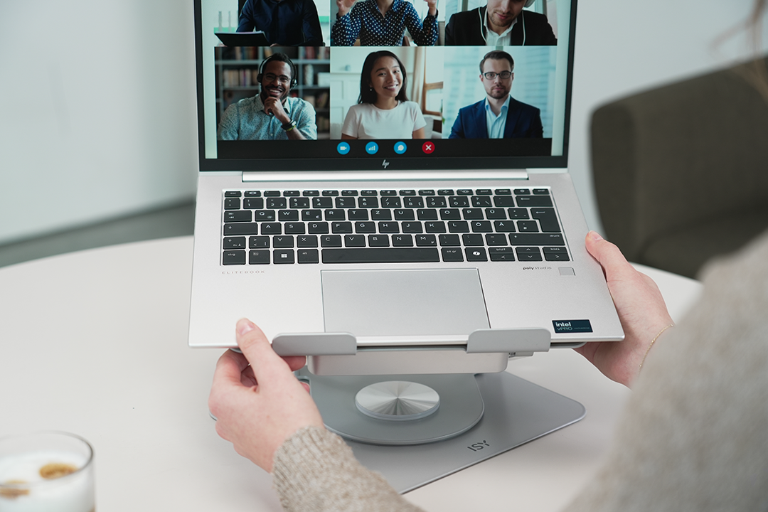 Egy személy egy nyitott laptopot tart egy állványon. A képernyőn egy videóhívás látható hat személlyel. Egy pohár itallal.