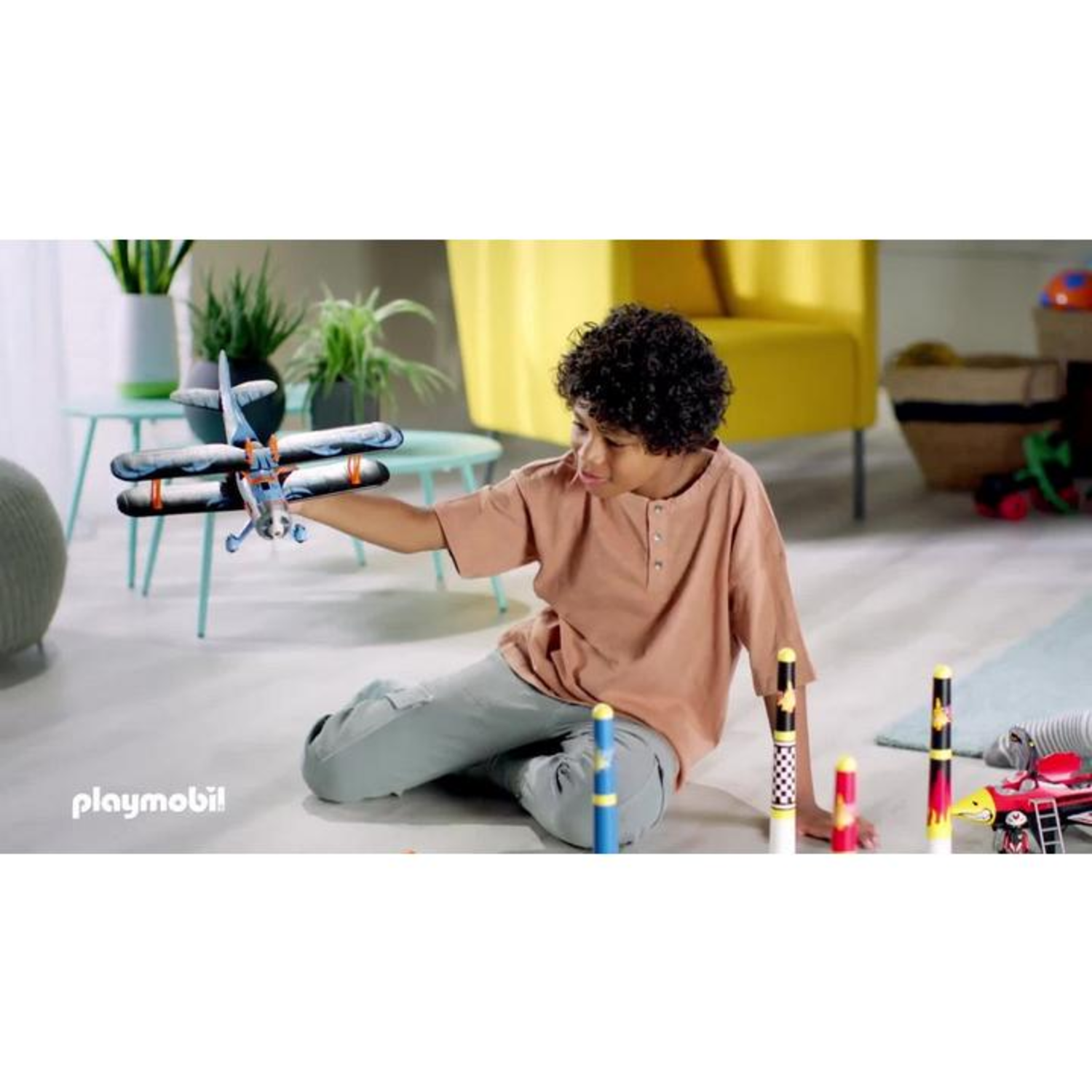 A boy holds a toy airplane. He sits on the floor. Various toys are on the floor. A yellow chair and potted plants are in the background.