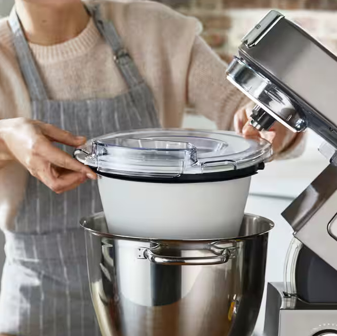 Osoba montuje mikser kuchenny. Umieszcza białą miskę z przezroczystą pokrywką w większej misce ze stali nierdzewnej.