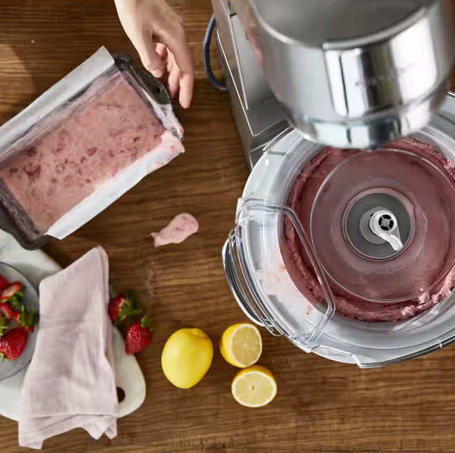 Osoba robi lody maszyną. Truskawki, cytryny i pojemnik z różowymi lodami na drewnianym stole.