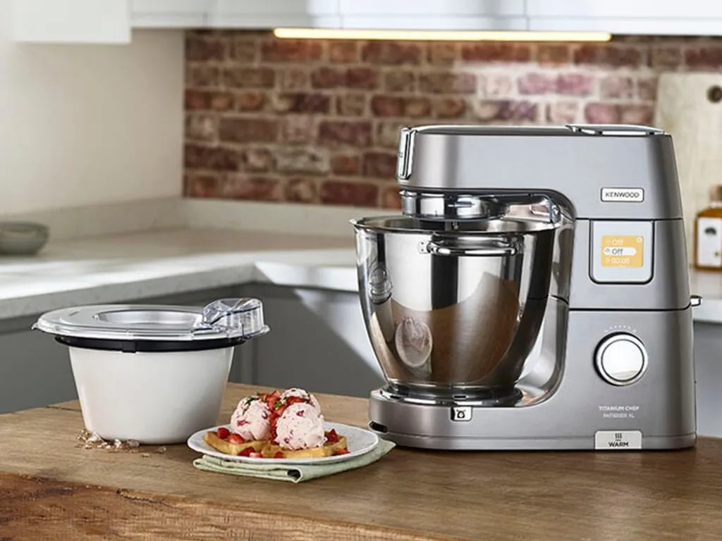 Srebrny mikser Kenwood obok miski i gofrów z lodami na drewnianym stole. Ceglana ściana w tle.