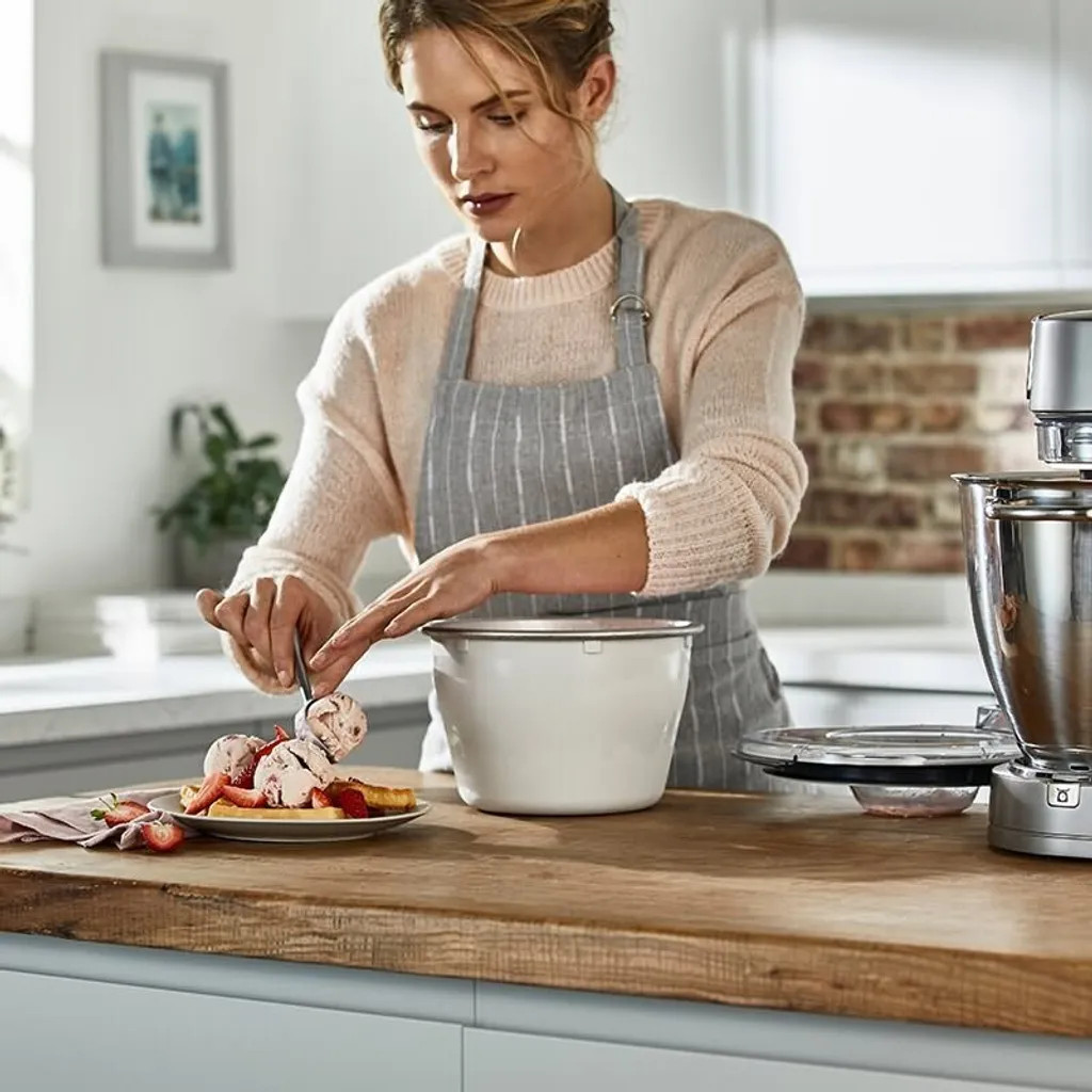 Kobieta w fartuchu nakłada lody na gofry z truskawkami na drewnianym blacie. Z prawej strony znajduje się mikser.