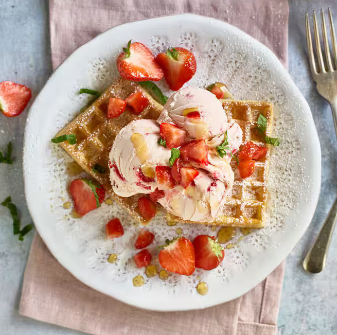 Talerz z goframi, lodami i truskawkami. Danie jest na różowej serwetce z widelcem i szarym tłem.