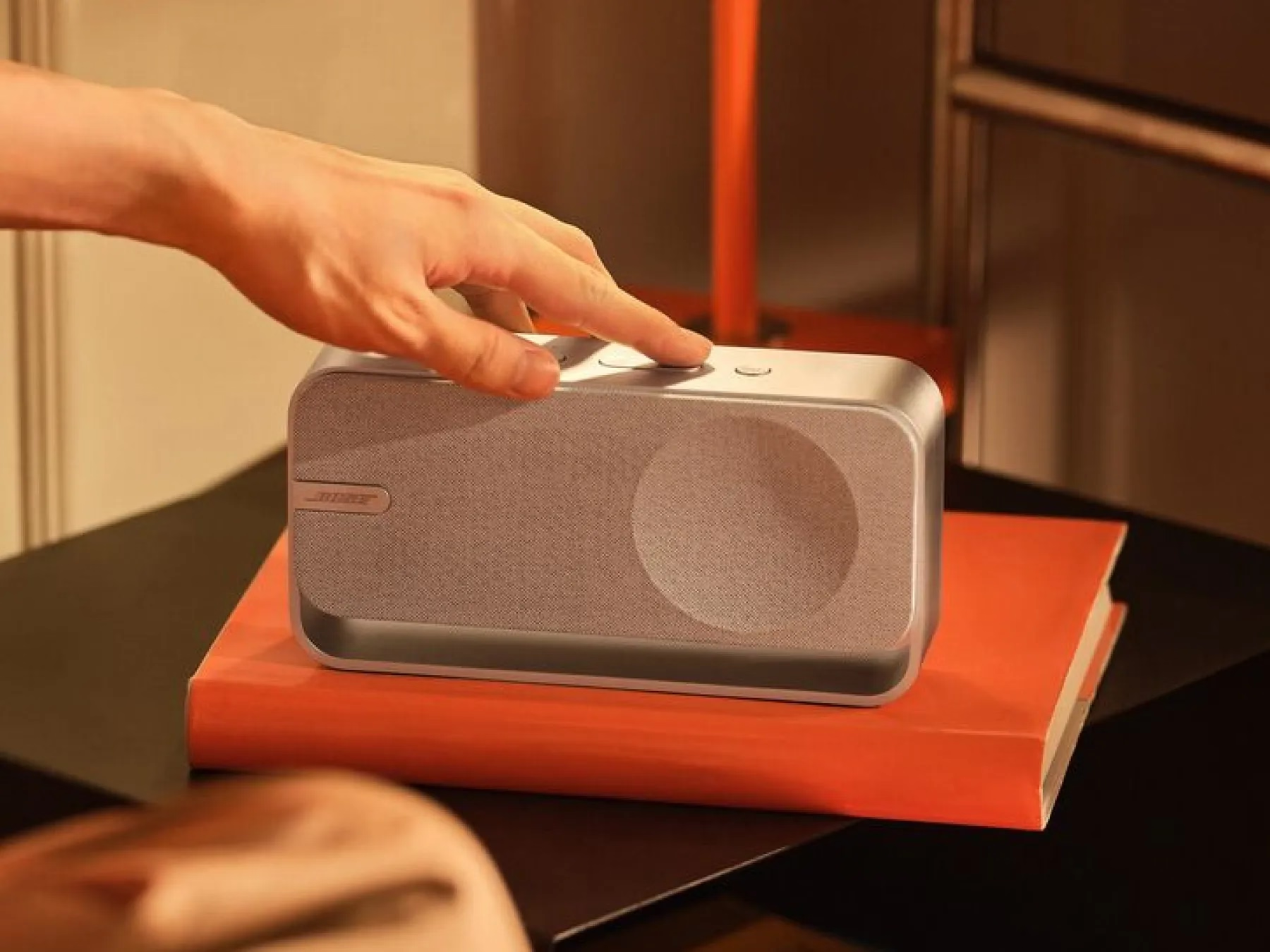 Eine Hand drückt eine Taste auf einem Bose-Lautsprecher, der auf einem orangefarbenen Buch liegt. Der Lautsprecher ist silber und grau. Der Hintergrund ist braun.