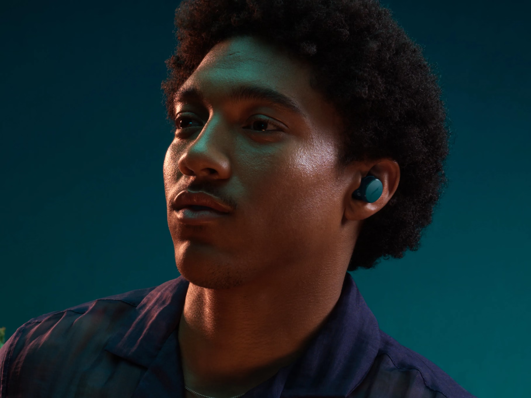 Un homme aux cheveux bouclés et aux écouteurs regarde vers la gauche. Il porte une chemise bleue sur un fond bleu-vert.