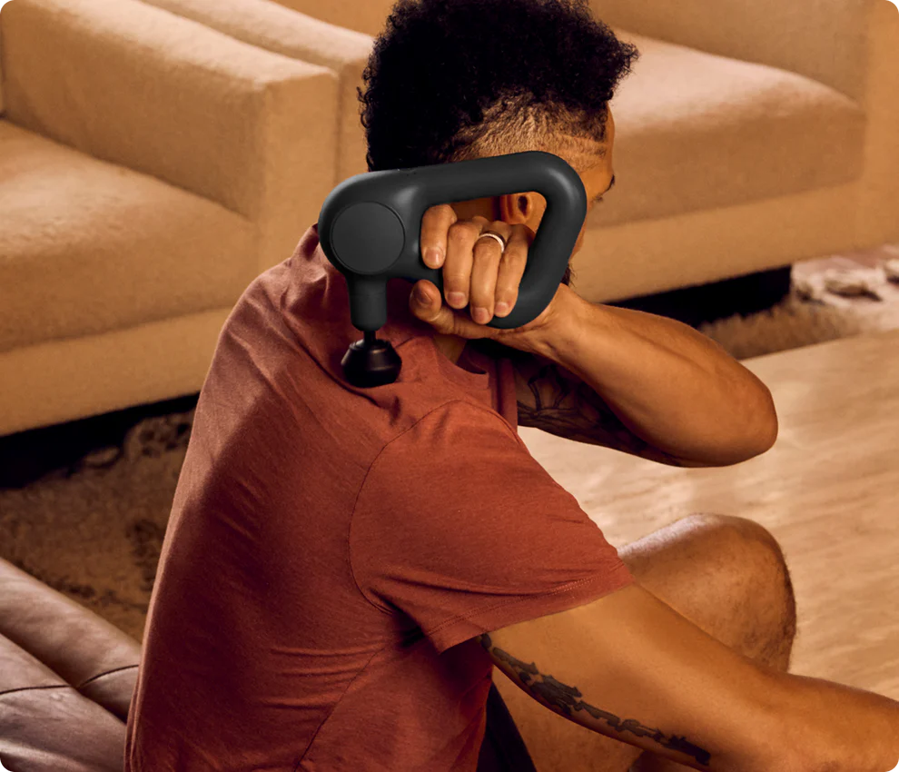 Ein Mann benutzt eine schwarze Massagepistole auf seiner Schulter. Er trägt ein rotes Hemd und hat ein Tattoo. Im Hintergrund ist ein Sofa.