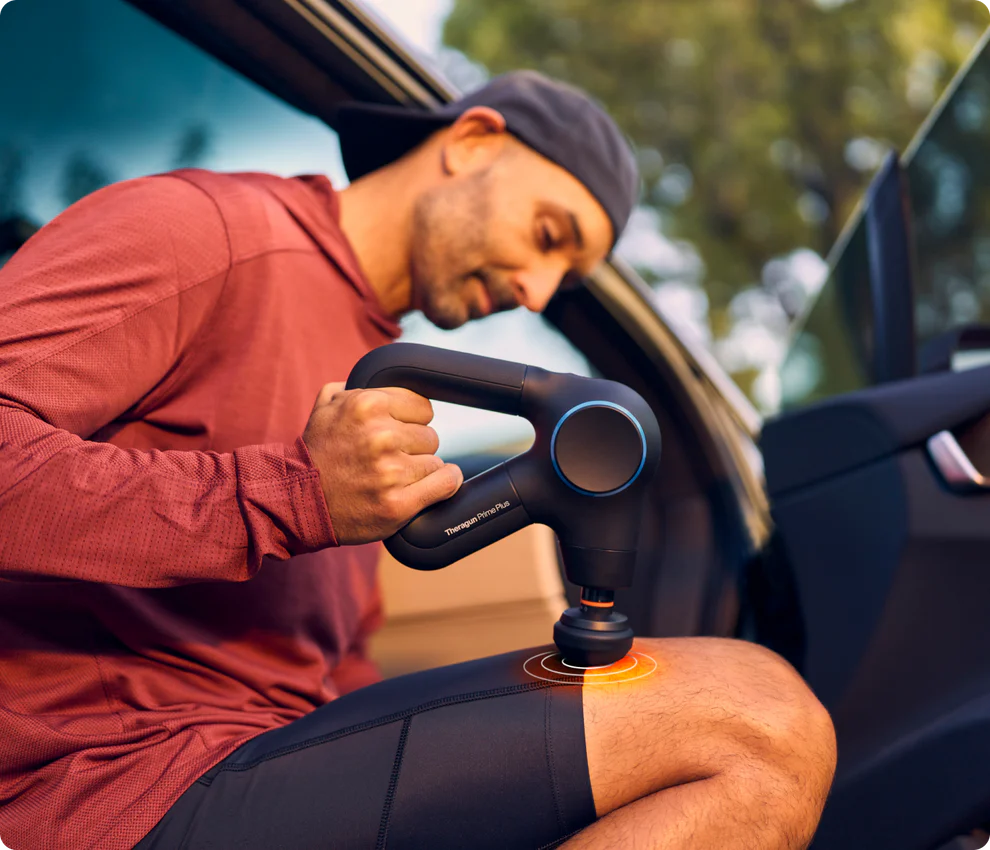 Mann benutzt Massagepistole an seinem Knie, sitzt im Auto. Die Pistole ist schwarz, Mann trägt rotes Hemd und Kappe.