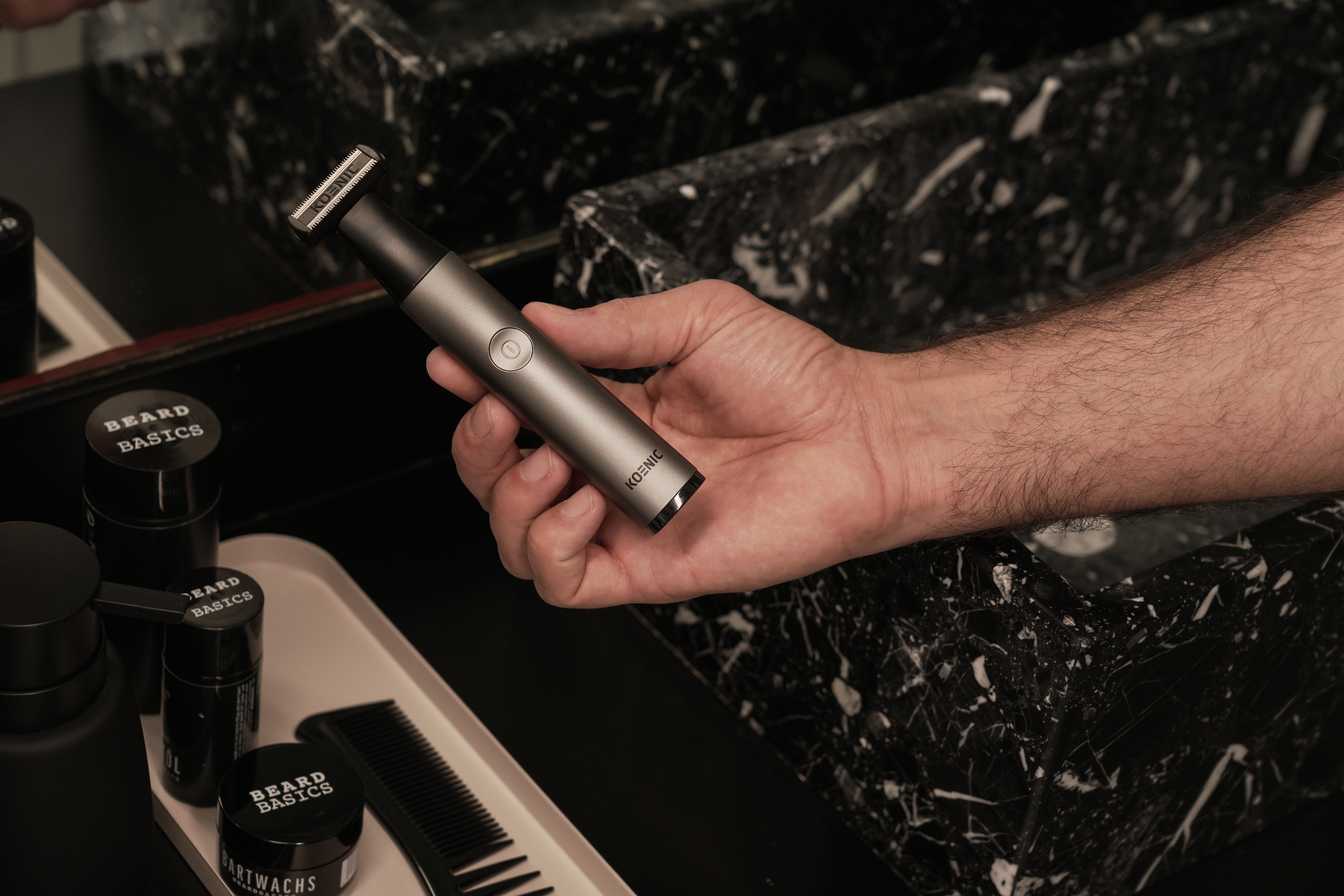 Ręka trzyma srebrną maszynkę do brody. Widoczne czarne pojemniki z napisem 'Beard Basics'. Tło to ciemna, marmurowa powierzchnia.