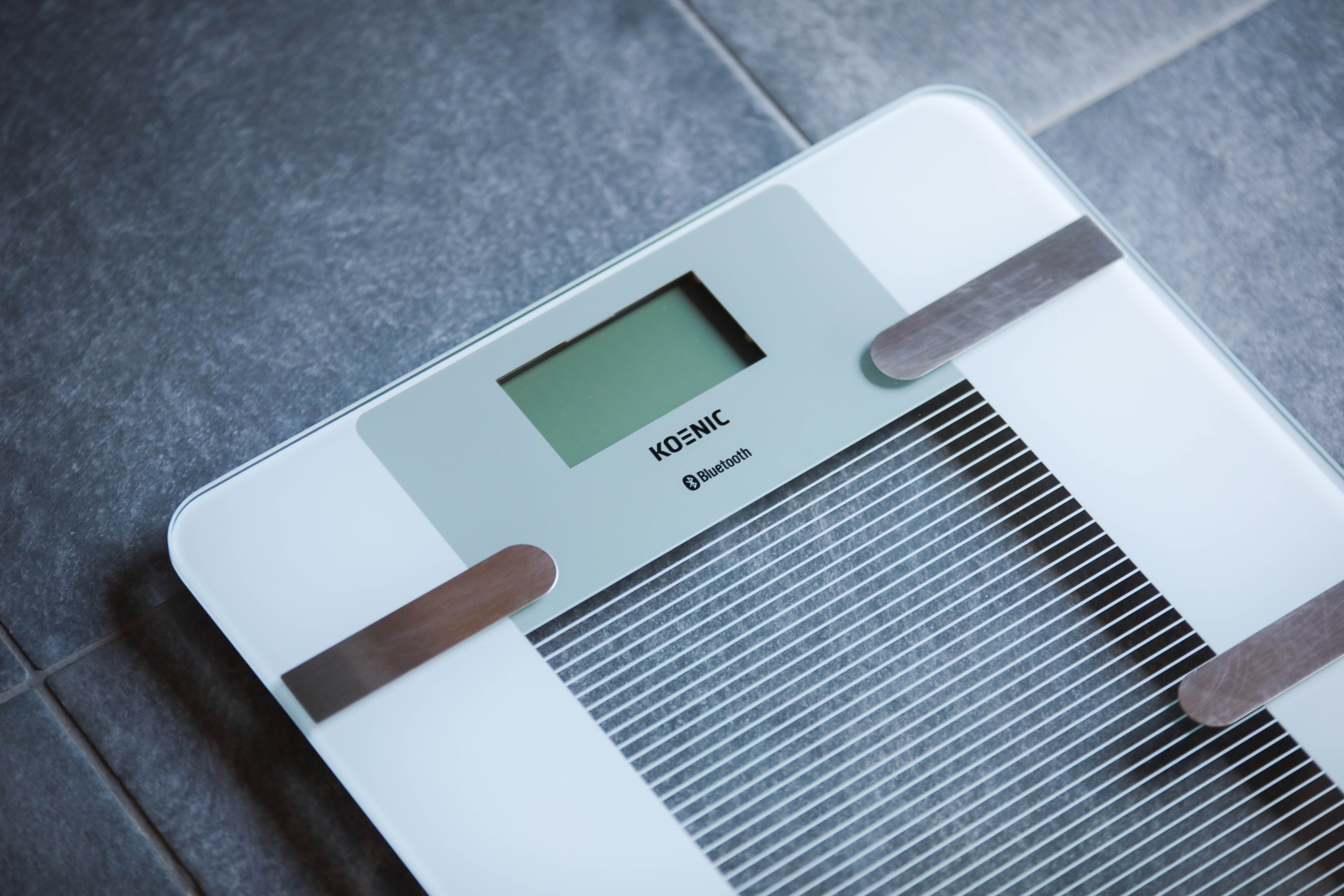 Biała i szara waga Koenic Bluetooth na szarej podłodze z płytek. Waga wyświetla zielony ekran i ma srebrne akcenty.