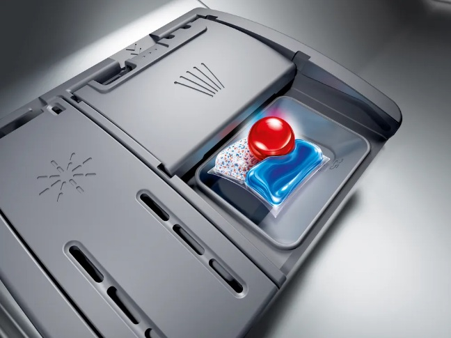 Przedział na detergent do zmywarki z trzema kapsułkami detergentu. Widoczne są czerwona, niebieska i biała kapsułka.