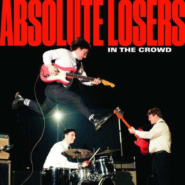 Band spielt auf der Bühne. Ein Gitarrist springt, während er eine rote Gitarre spielt. Schlagzeuger und Bassist spielen ebenfalls. Text sagt 'Absolute Losers'.