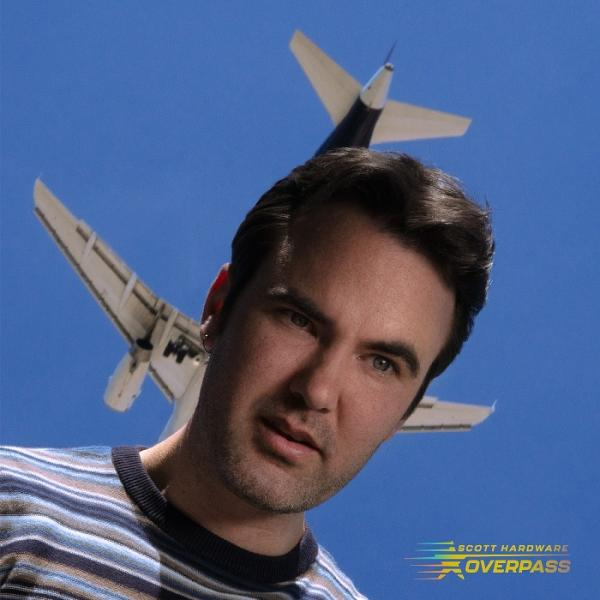 Ein Mann blickt den Betrachter an, ein Flugzeug ist hinter ihm, auf blauem Himmel. Er trägt ein gestreiftes Hemd.
