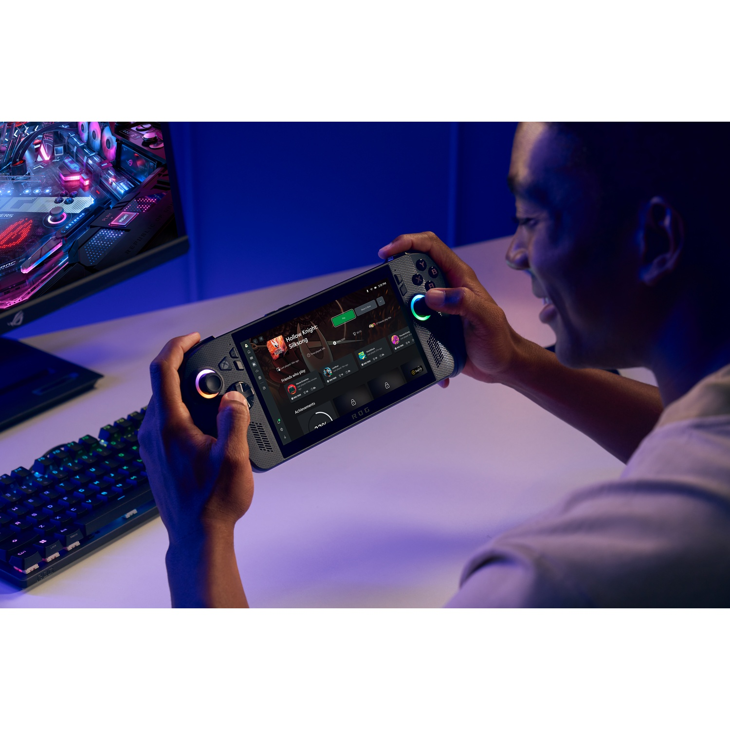Una persona tiene in mano un dispositivo di gioco portatile, che gioca. Sullo sfondo, una tastiera e un monitor con illuminazione colorata.